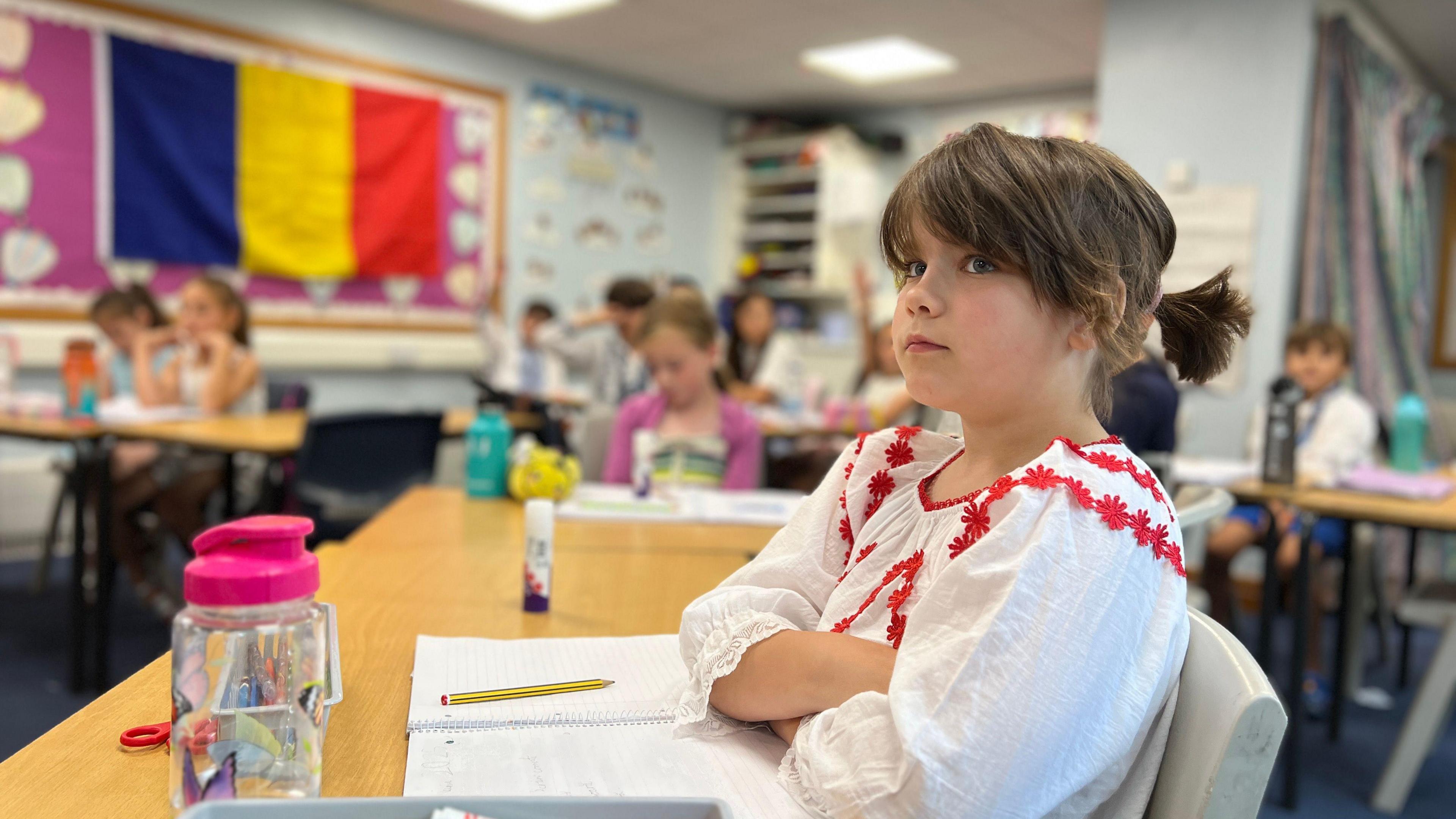 A classroom scene with students seated at desks working on assignments. In the foreground, a student wearing a white shirt with red embroidery sits with arms crossed. The background includes other students, a large flag on the wall, and desks scattered with school supplies like notebooks, pencils, and a basket of markers