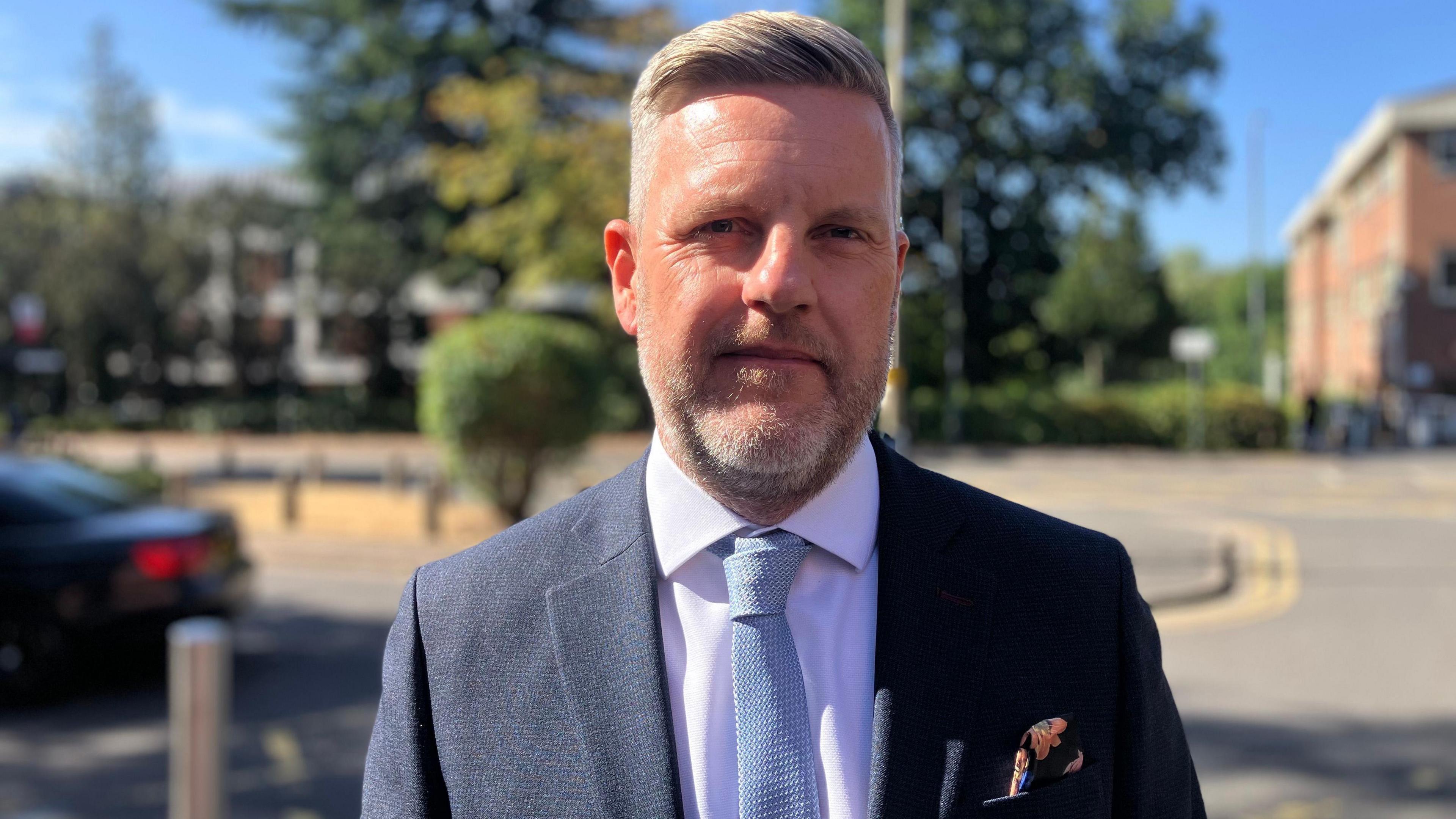 A man with grey hair styled to the side. He is wearing a white shirt, light blue tie and black suit jacket with a navy and pink patterned handkerchief in the breast pocket