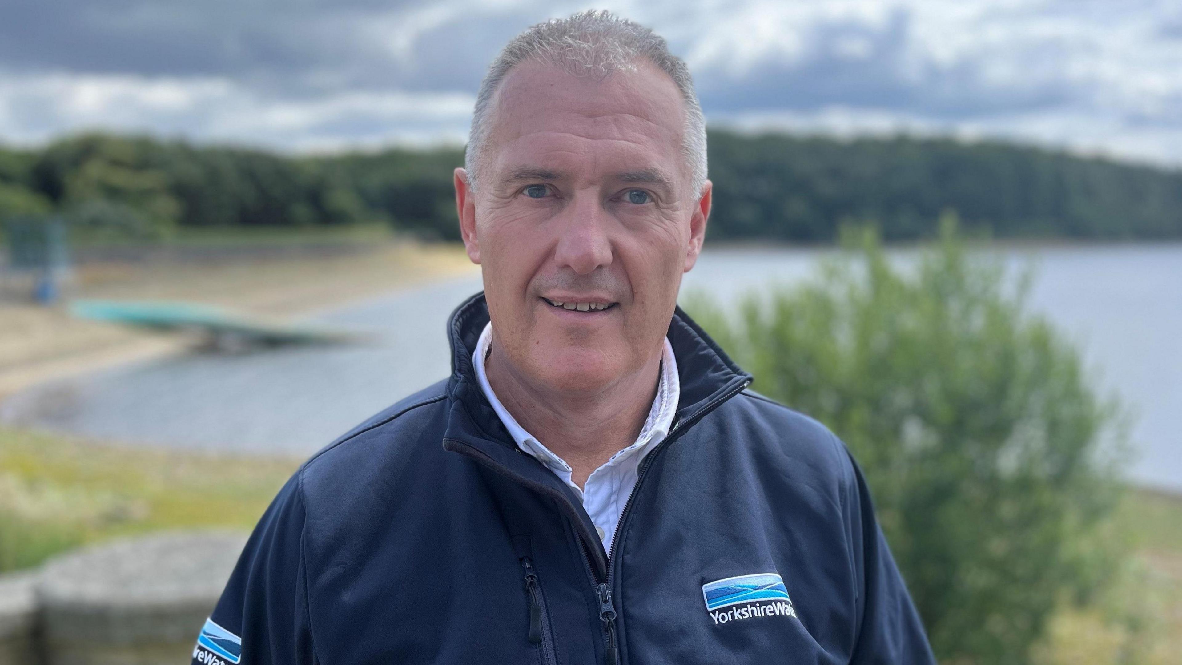 Dave Kaye, who has short grey hair and blue eyes. He is wearing a blue Yorkshire Water zip-up jacket and he is standing in front of a reservoir.
