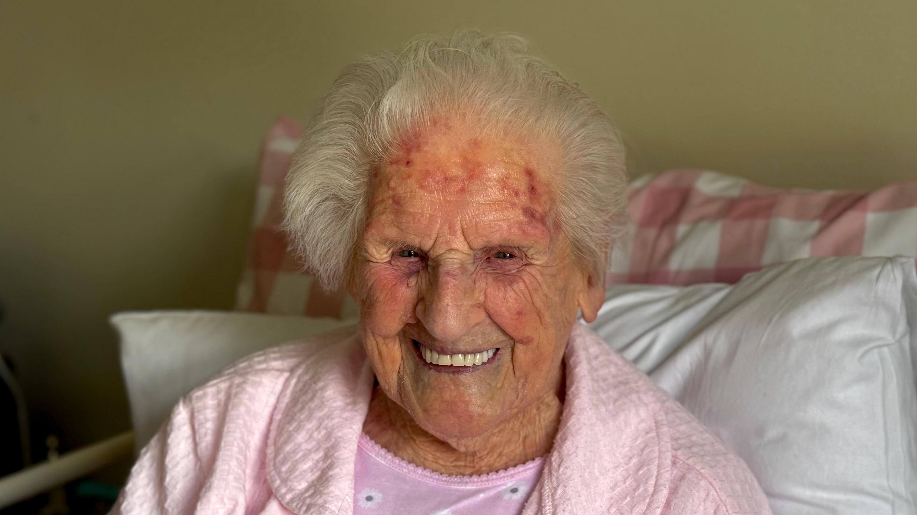 Elderly woman with white hair is smiling at the camera. She is wearing a pink dressing gown.