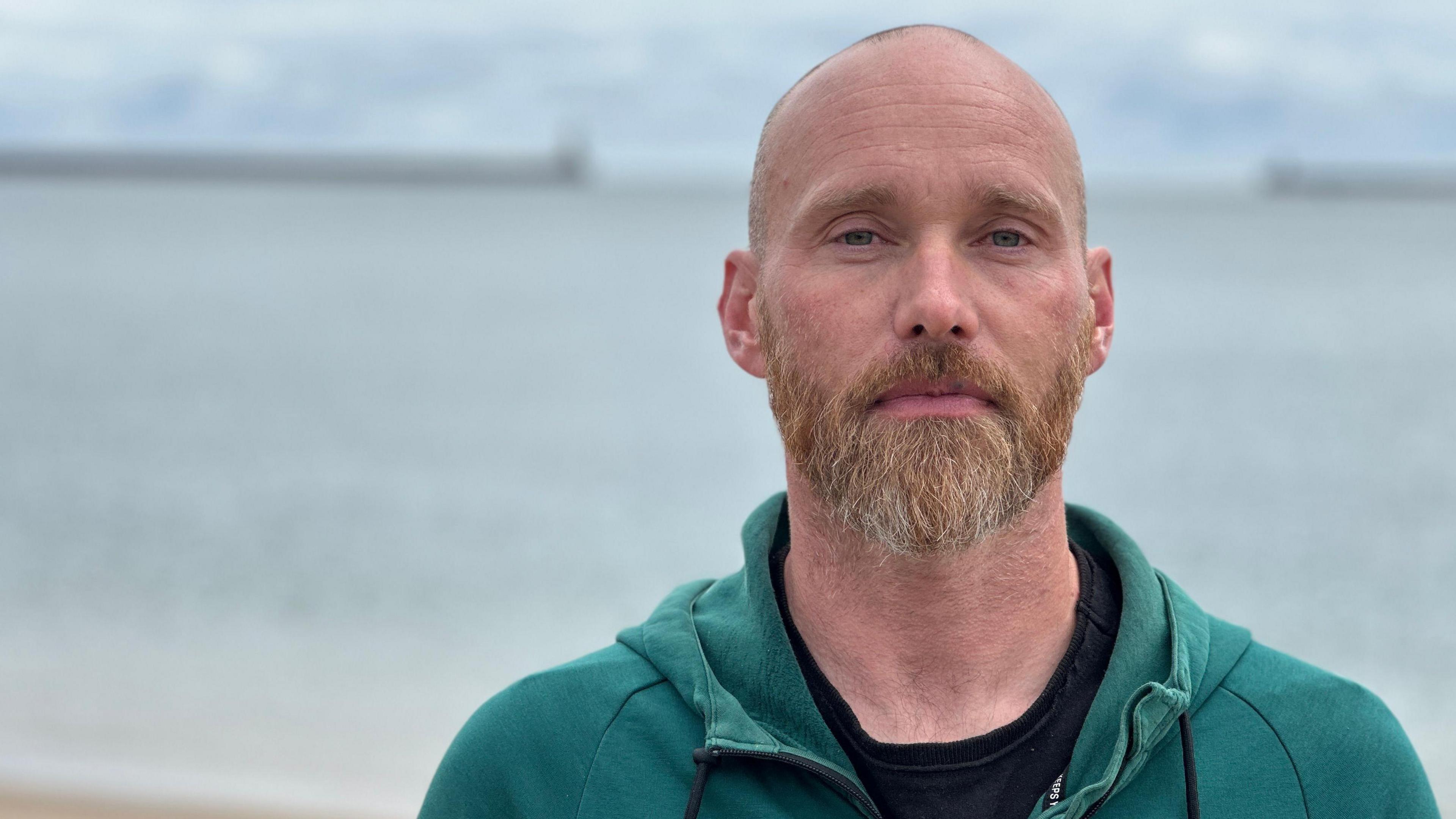 Olly Armstrong is standing with his back to the sea on a beach. He is mostly bald and has a thick beard and moustache. In the background, which is out of focus, is a pier with a lighthouse on it. He is wearing a teal coloured hoodie and black t-shirt.
