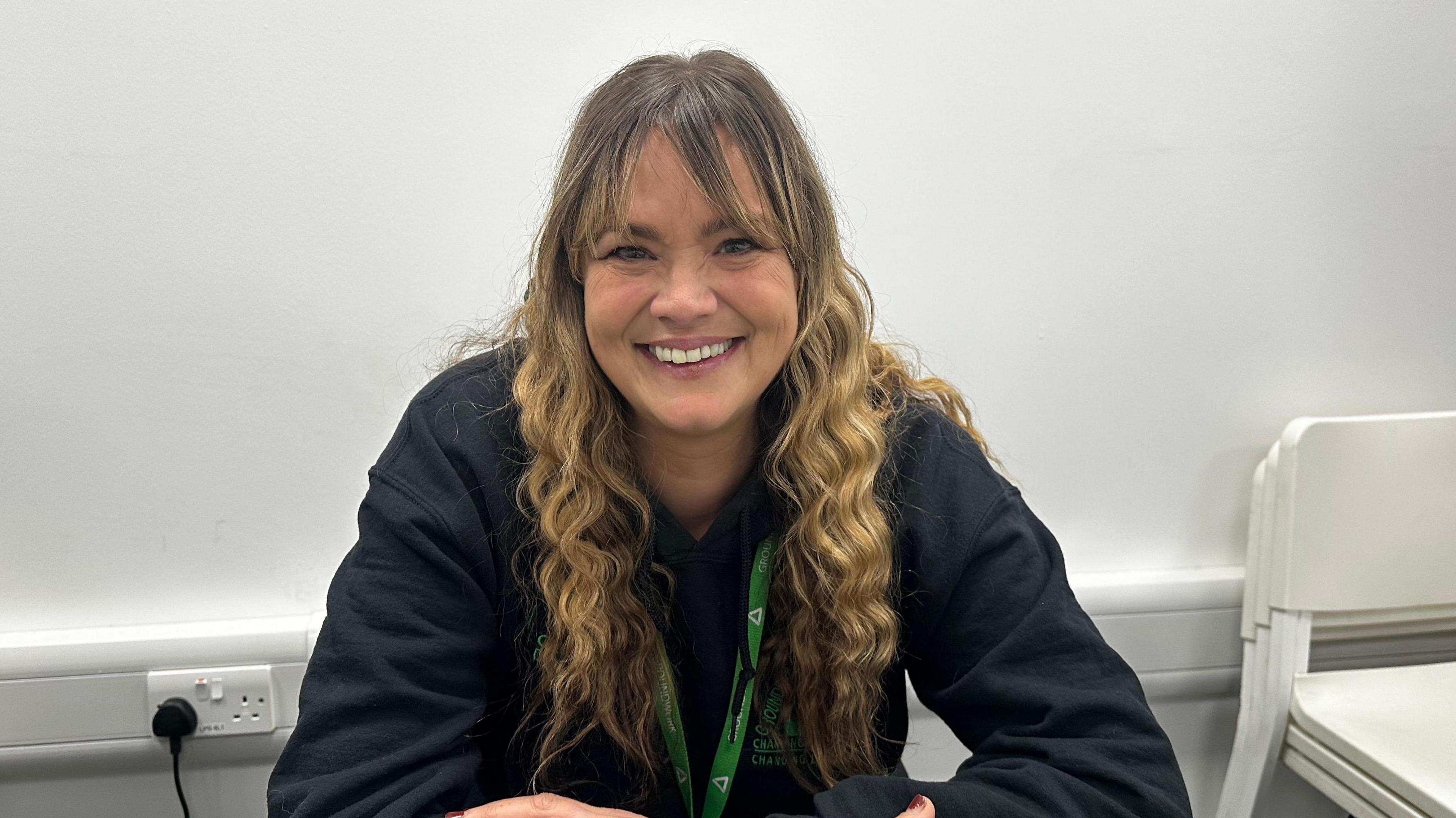 A woman wearing a black jacket and green lanyard smiles at the camera in front of a white wall