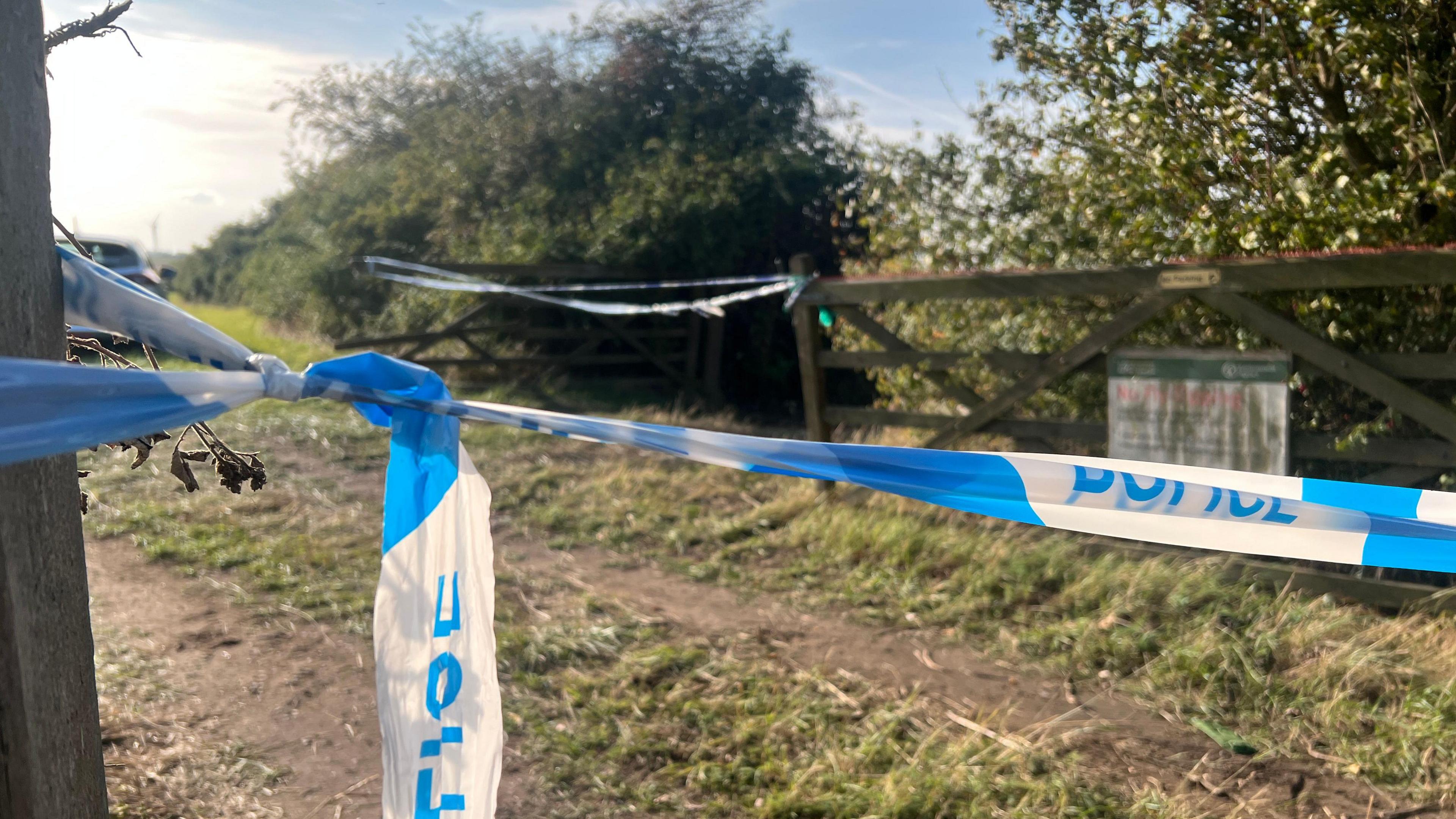 Blue and white police tape stretches across an entrance into a field. More tape can be seen ahead across other gates into the field.
