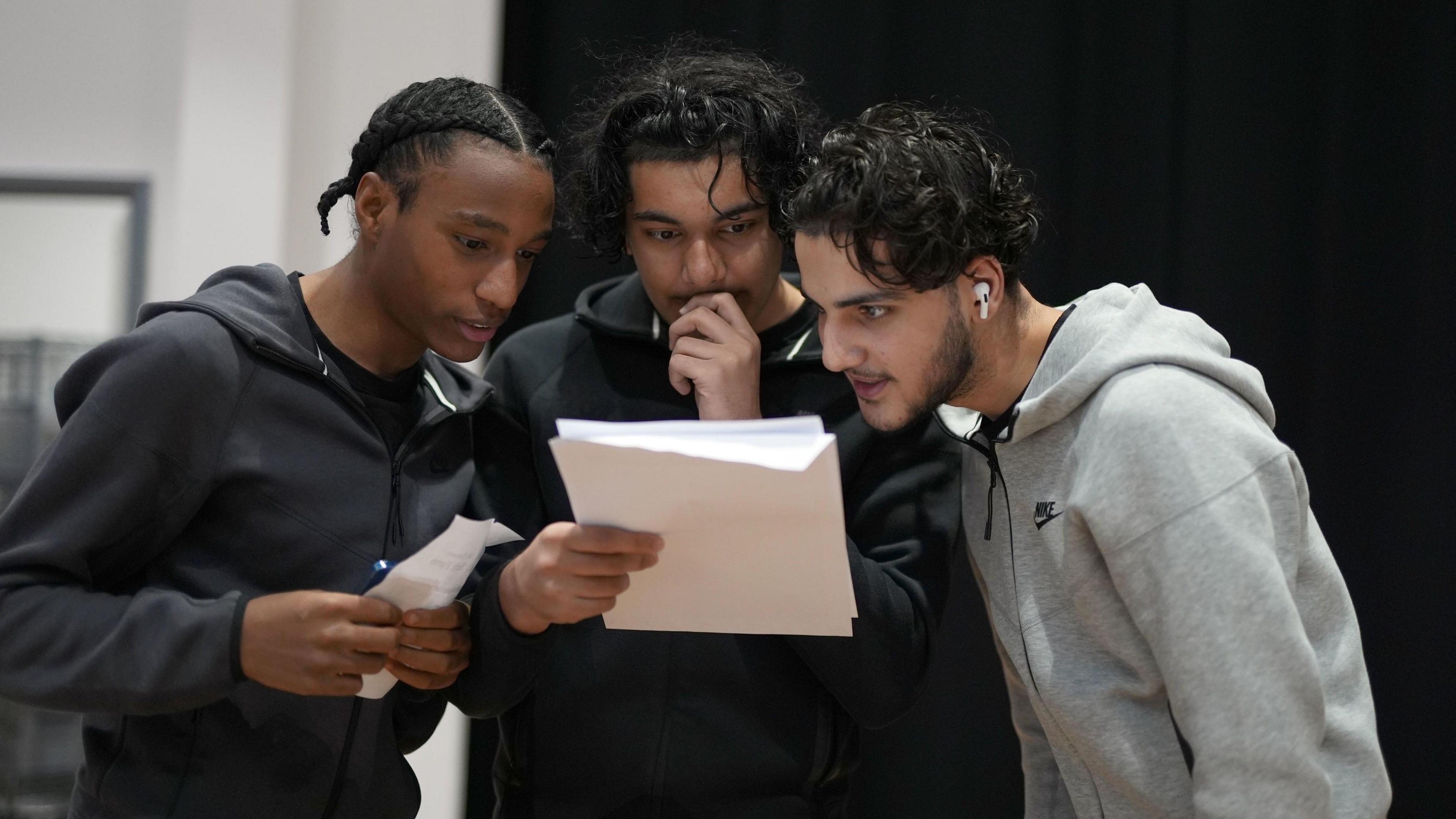 Three young men anxiously read the one in the middle's A-level results. He is holding his hand up to his mouth.