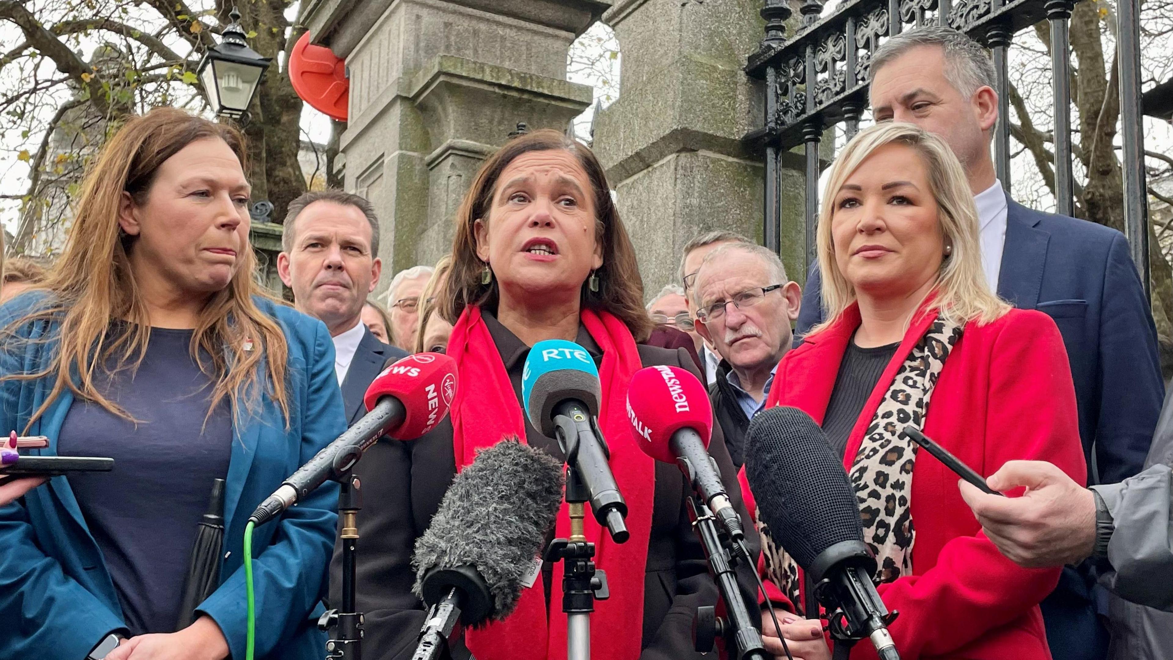 Sinn Fein President Mary Lou McDonald (centre) and Vice President Michelle O'Neill speak to the media on Merrion Square in Dublin. Mary Lou has shirt brown hair, and wearing a black coat and red scarf speaking into microphones. Michelle has shoulder length blonde hair and wearing a red coat and leopard print scarf.