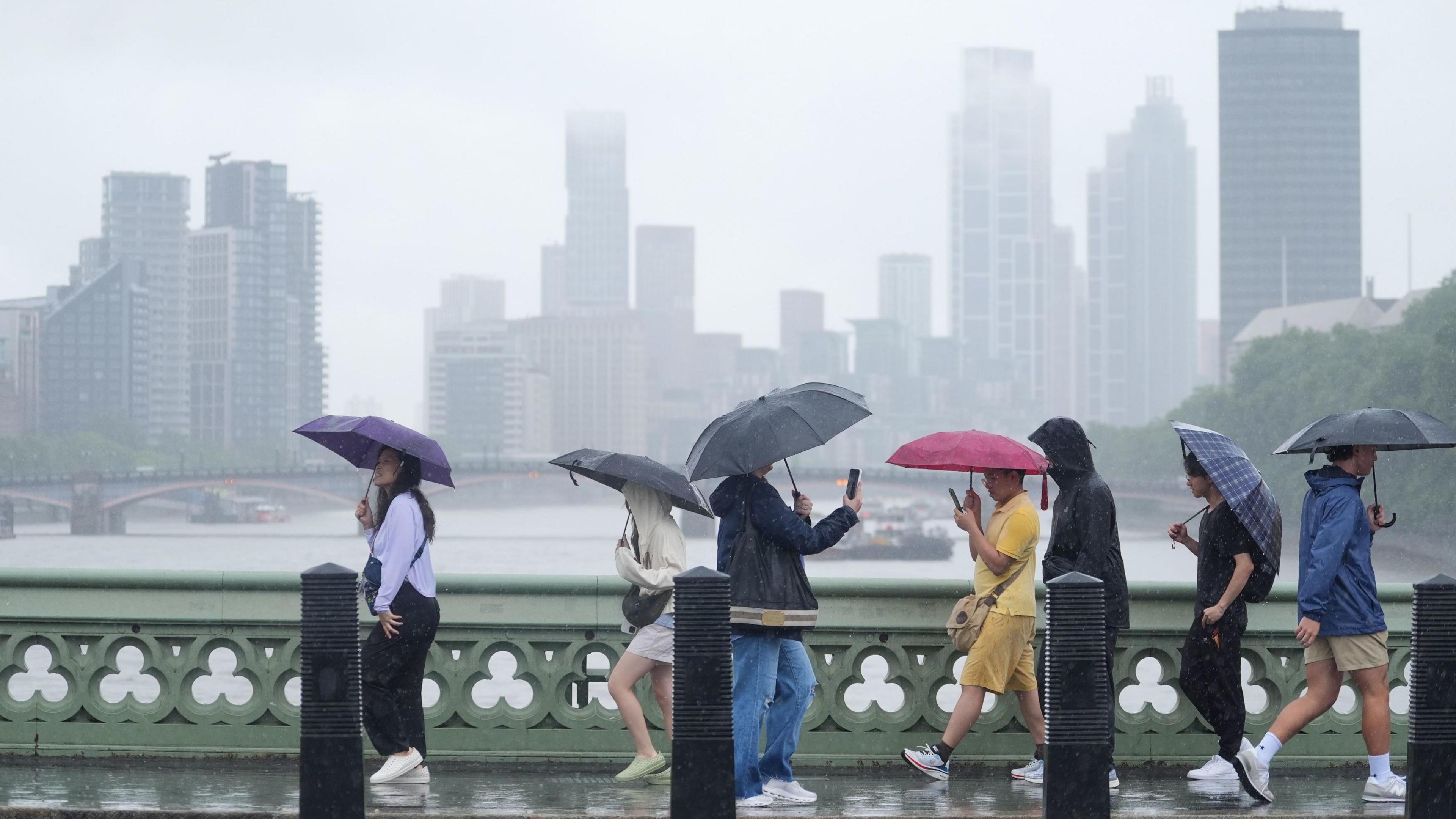People with umbrellas walk over a bridge in a rain storm with skyscrapers and a river and another bridge in the background