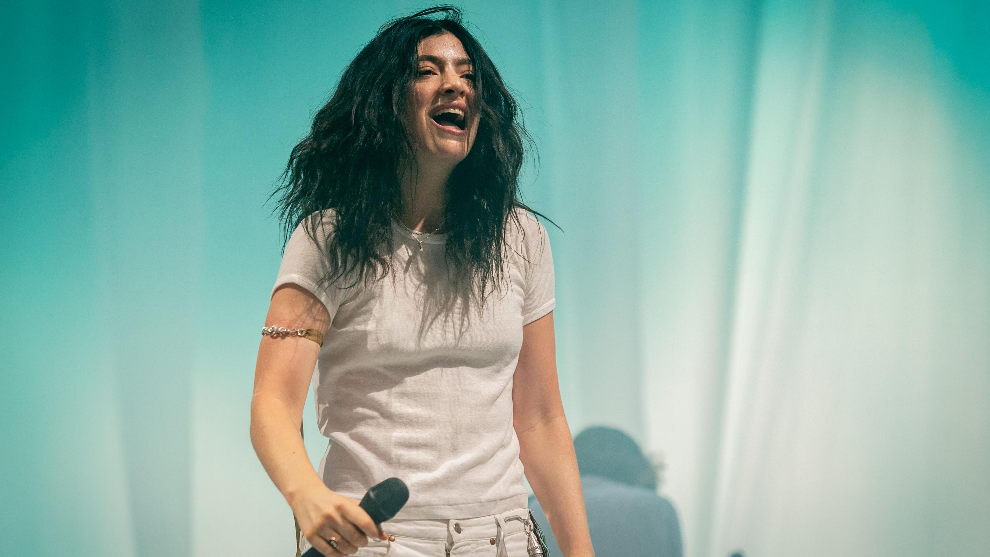 Singer Lorde wears a white t-shirt and trousers in front of a turquoise curtain on-stage. She's holding a microphone and smiling.