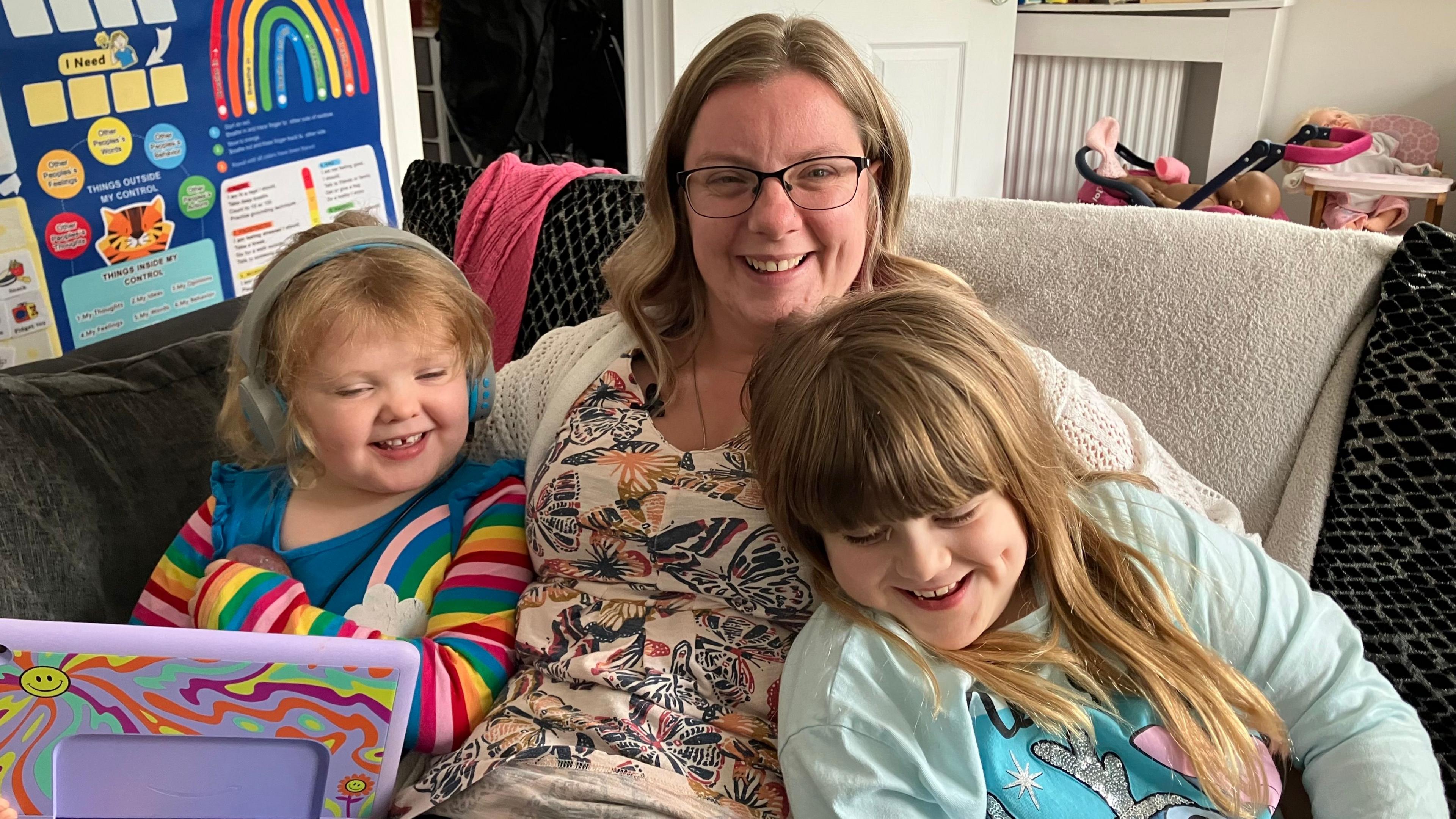 Hayley sitting on the sofa in her living room with her arms around her two daughters. They are all smiling and playing, wearing colourful clothes. Hayley has long way strawberry blond hair and glasses and is wearing a beige blouse with butterflies on it.
