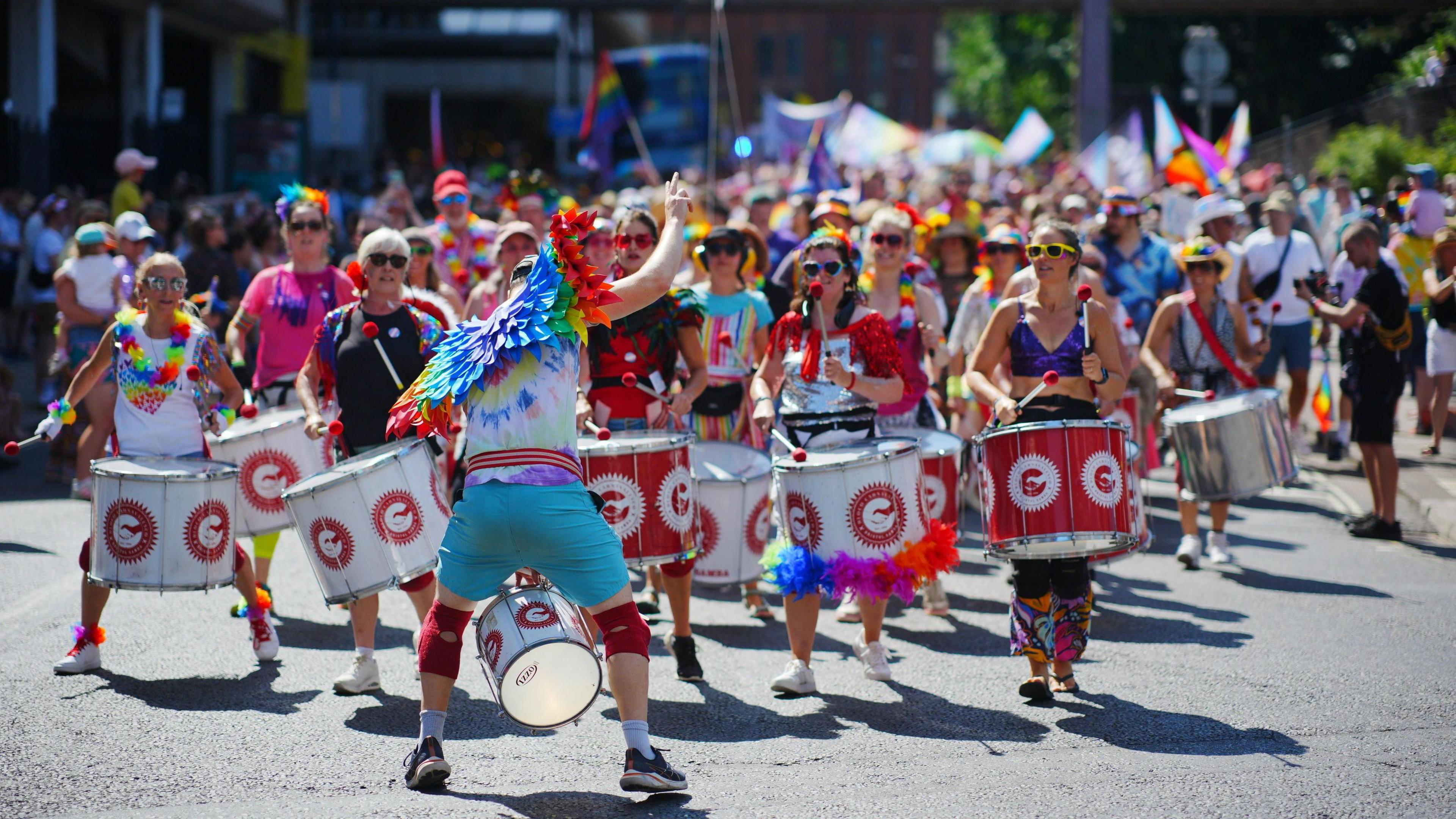 A shot looking down a large pride parade - in the foreground are a samba band, all holding red and white drums and dressed for a summer's day with rainbow accessories. The leader has his back to the camera to face and instruct the band, and wears a pair of rainbow wings
