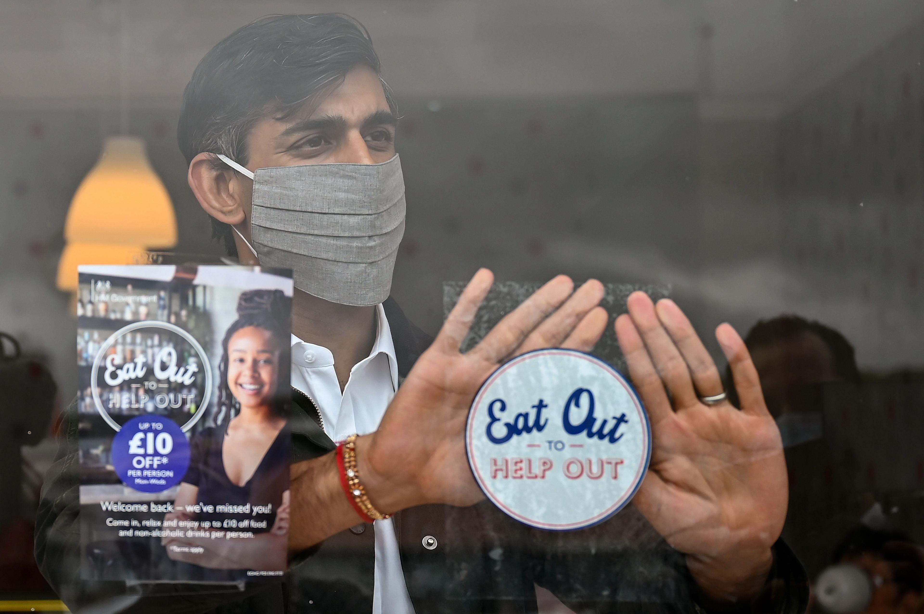 Photograph of Rishi Sunak wearing a mask in the summer of 2020. He is behind a window of a restaurant and attaching a sticker which promotes the Treasury's Eat Out to Help Out scheme.