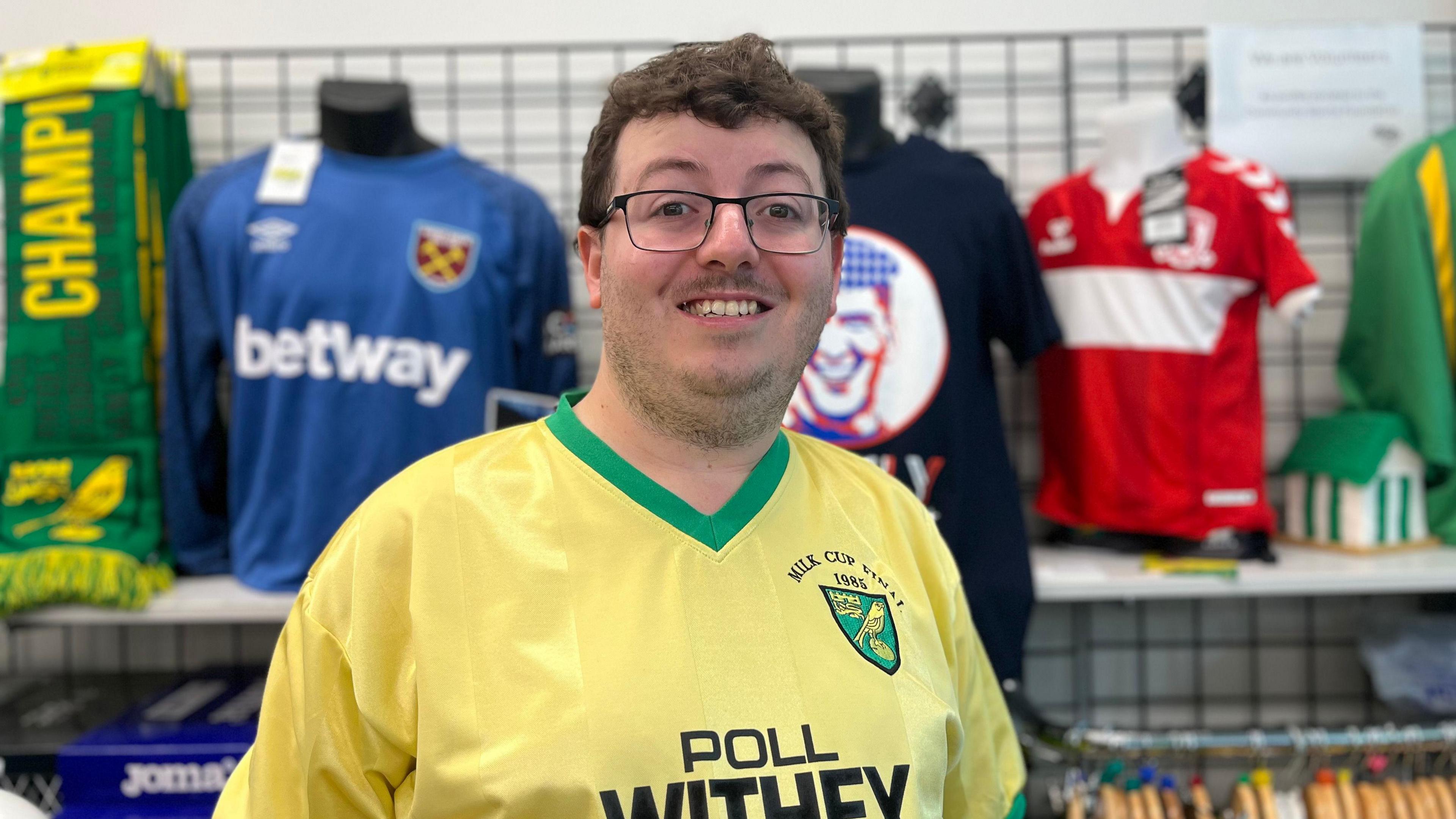 A picture of Jacob Bowles. He is wearing glasses and a retro yellow Norwich City football top and smiling. Other football shirts can be seen behind him.