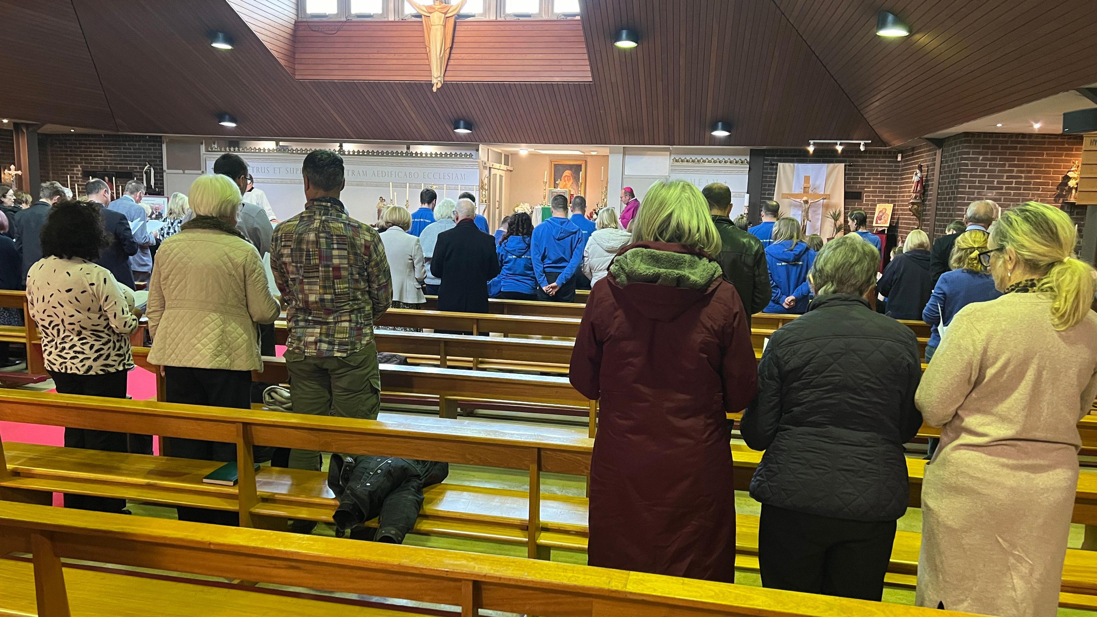 People dressed in coats in a church. They are standing with heads slightly bowed while a bishop in pink robes can be seen at the front of the church.