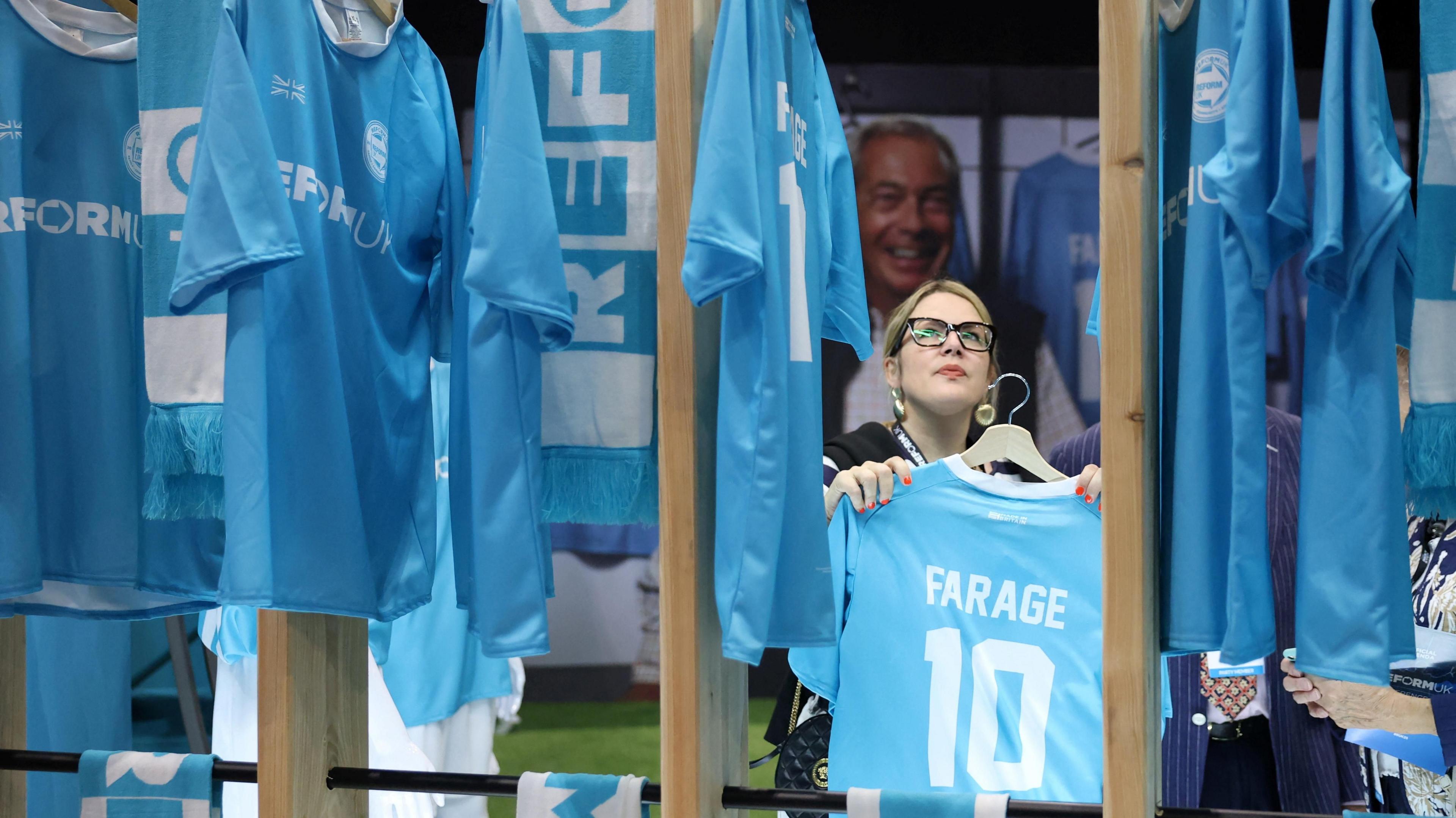 A woman hangs up pale blue football shirts, with Farage 10 on the back of them, at the Reform conference