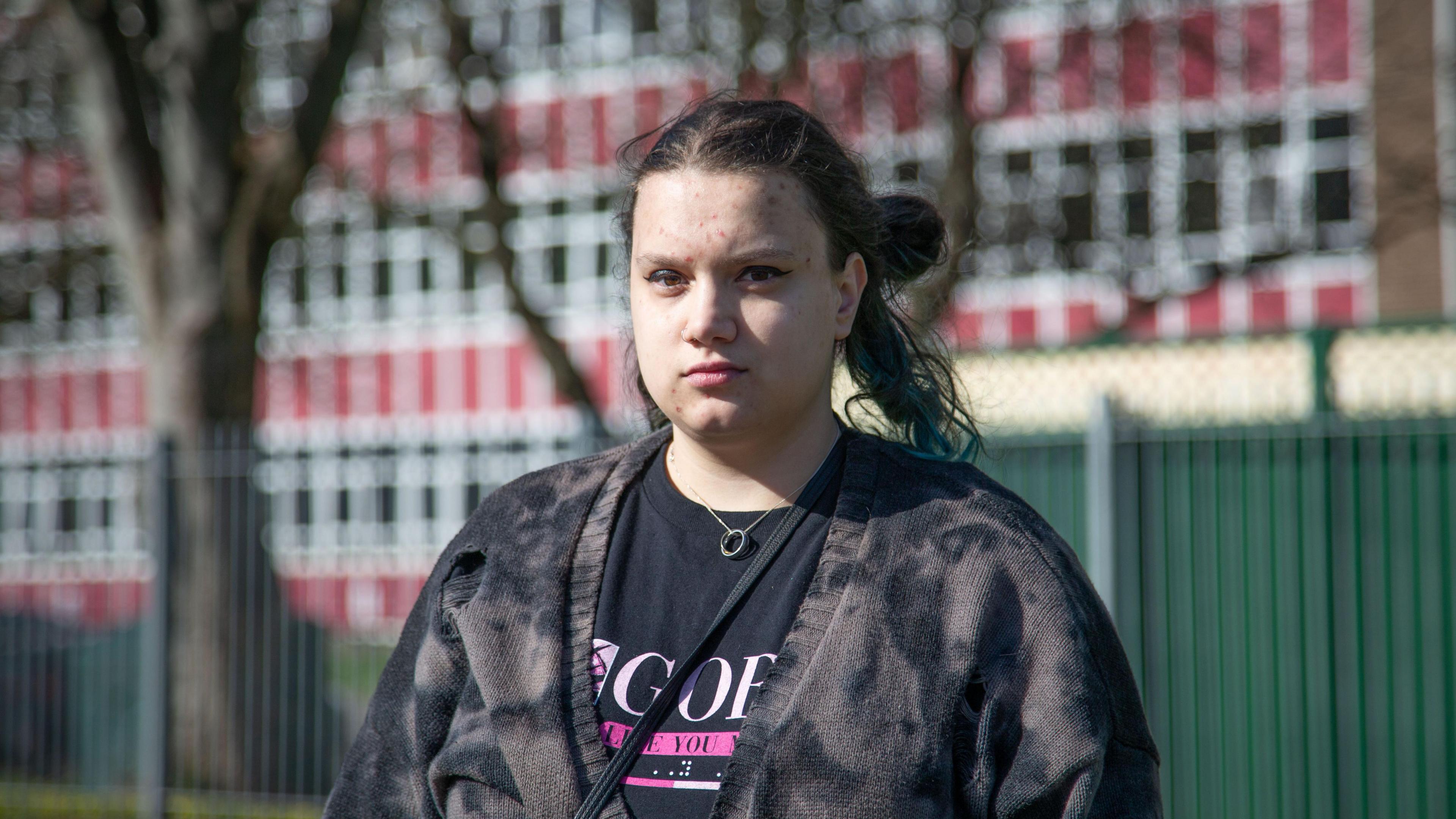 A young woman, with long dark hair tied up in a bun looks at the camera from outside a large school building. She wears a dark tie-dye cardigan and black t-shirt.