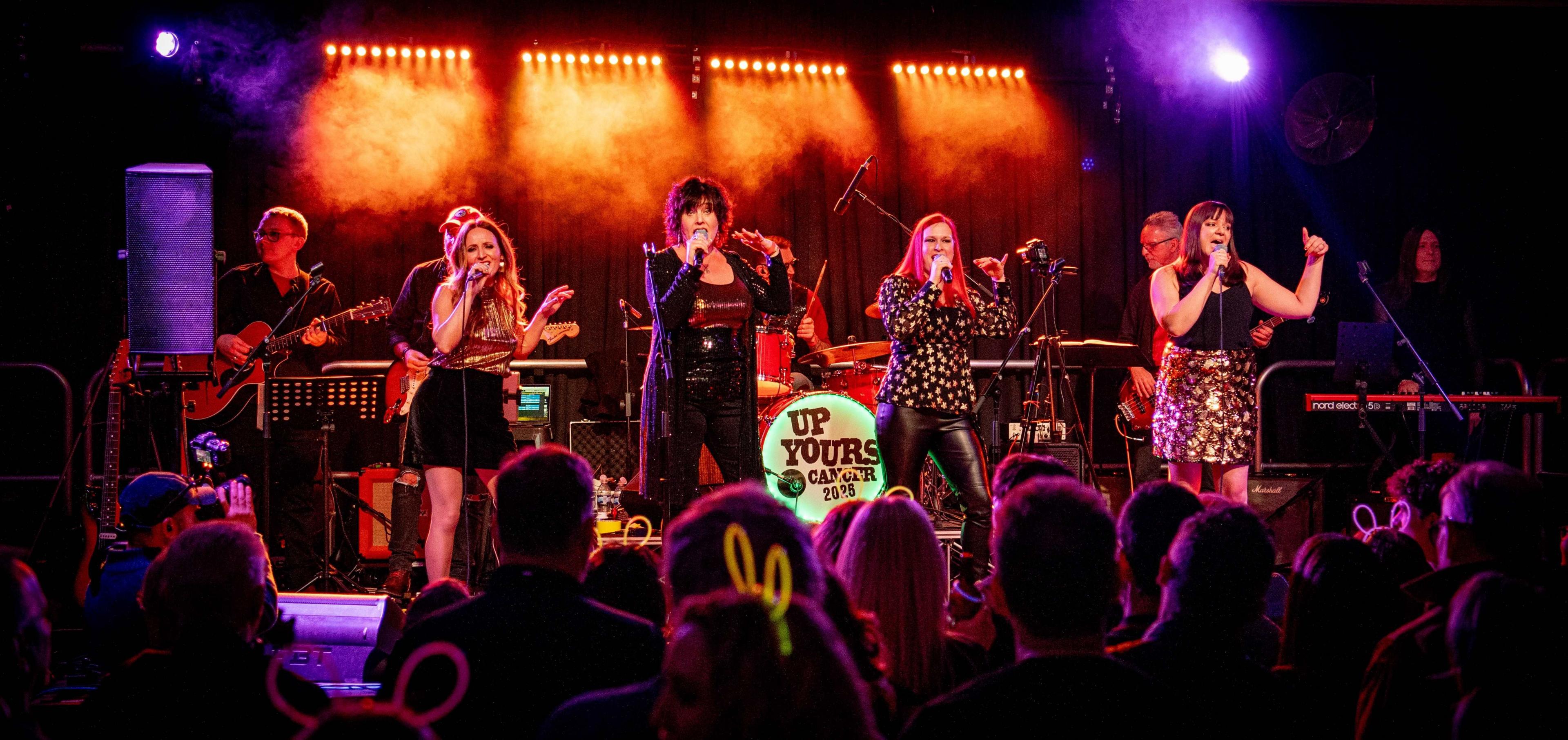 Four women in a line on stage. They are wearing gold and black outfits. Behind them are musicians and in front is the audience.