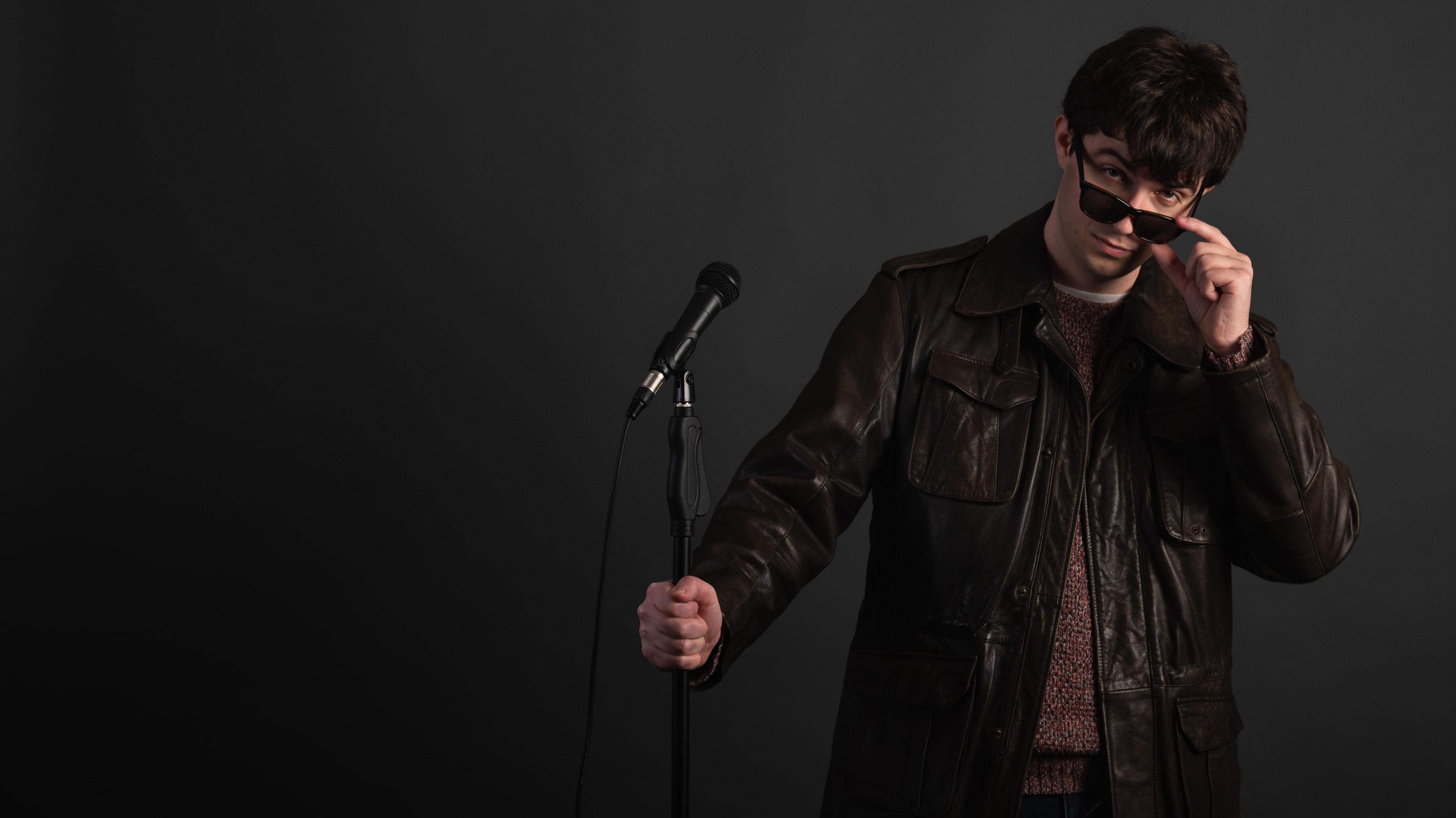 Burt Williamson wearing a black leather jacket and holding a mic stand in one hand, while pulling his sunglasses down from his eyes with the other hand.