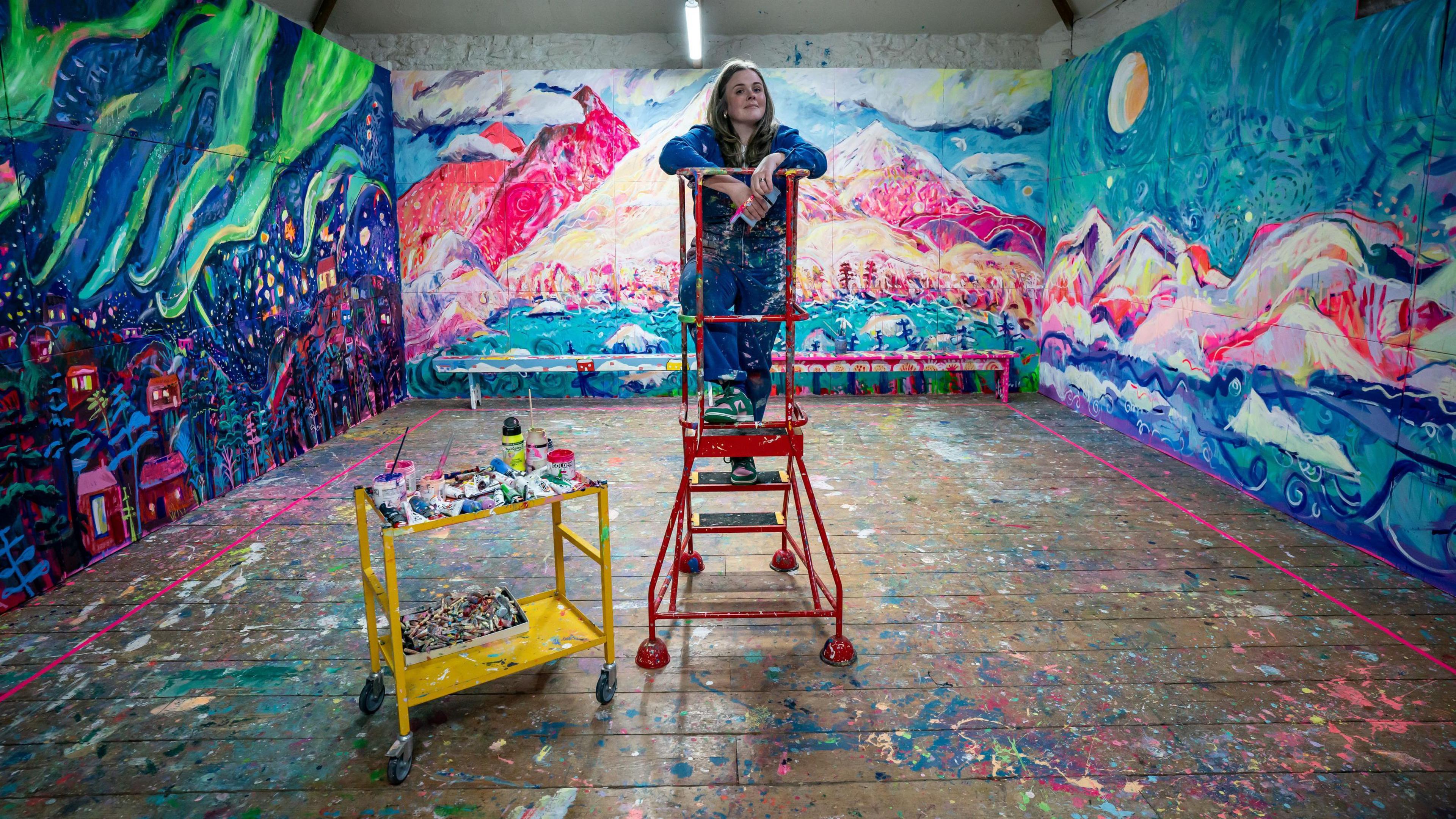 Artist Emily Powell stands on a red step ladder next to a yellow trolley with her paints on them surrounded by her work at a barn in Dartmoor.