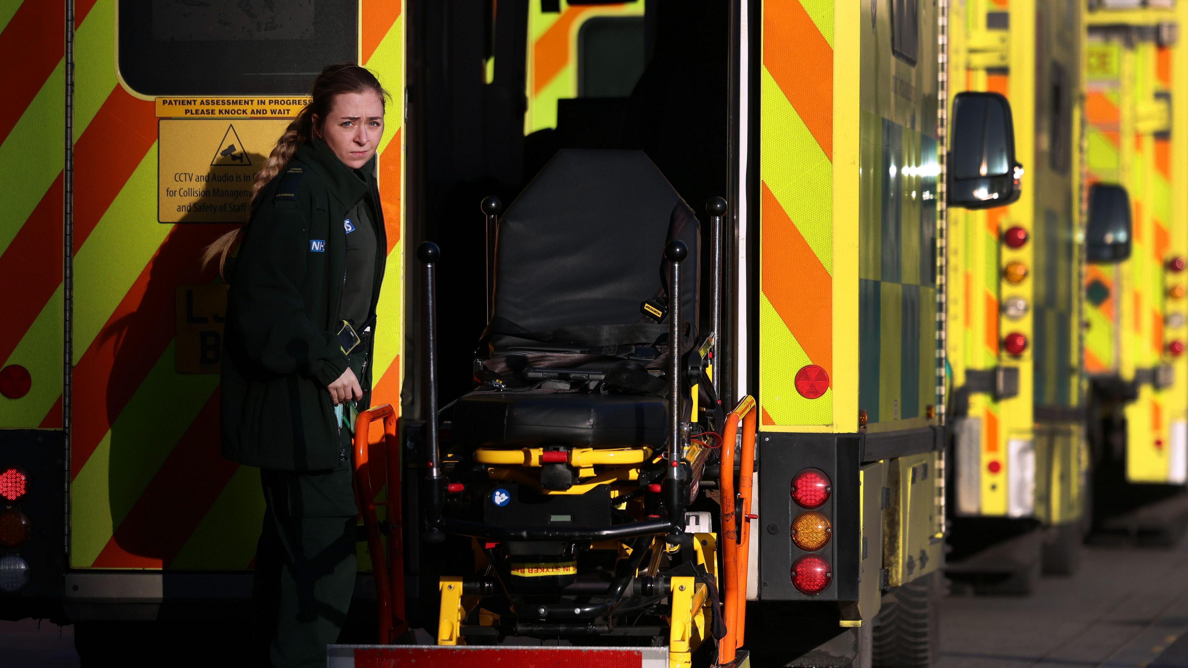 A female paramedic stands outside a yellow London Ambulance, holding a stretcher platform extended from the vehicle. Behind her, a line of ambulances are parked.