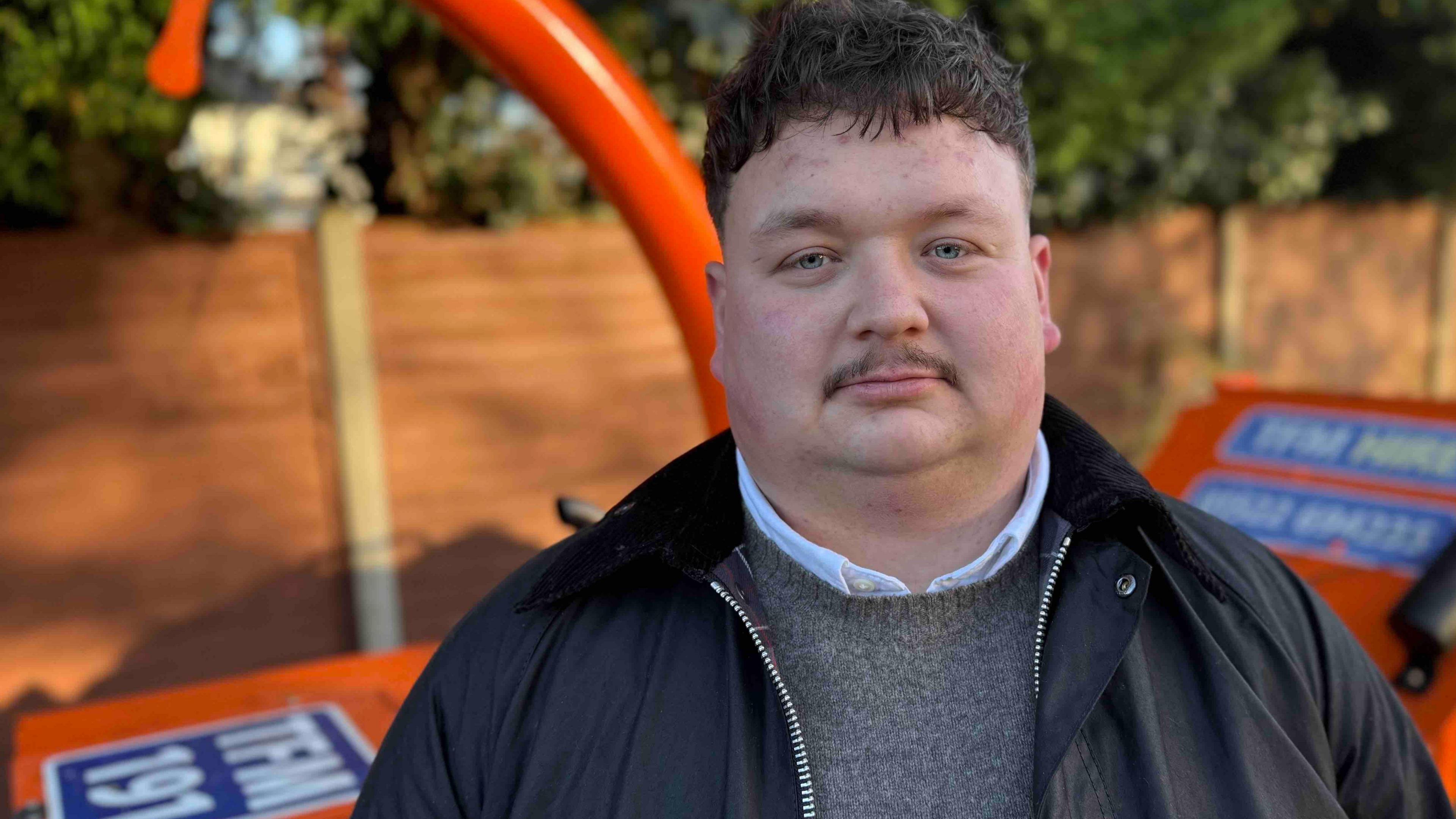 A man with a mop of black hair and a neatly trimmed moustache stands in front of a wood chipper - a large orange and blue machine with an orange arm rising high into the air. Behind it, out of focus, is a wooden fence and green trees.