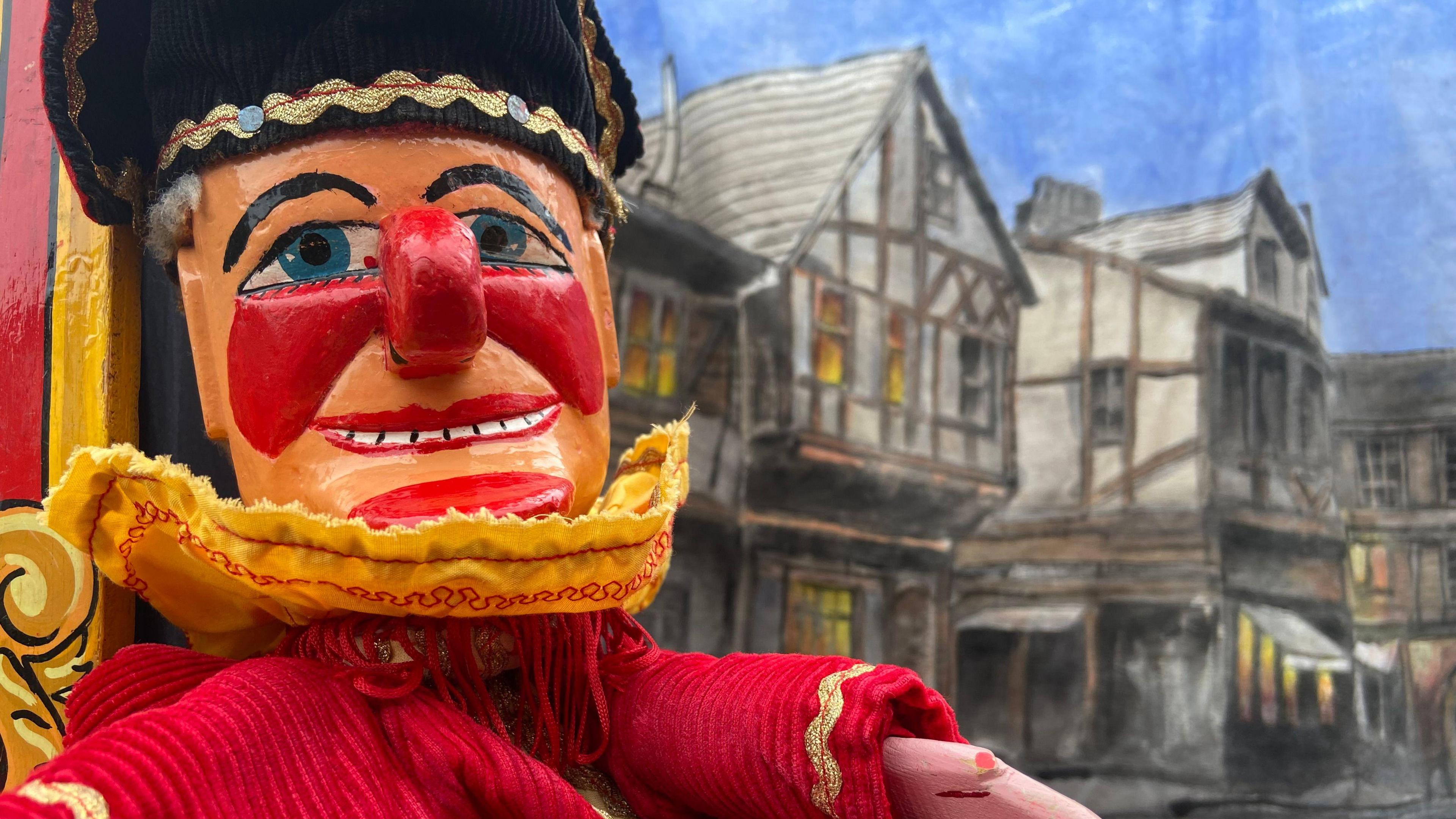 A traditional Mr Punch puppet, with a bright red nose, cheeks and chin, in front of a painted backdrop of Tudor houses.