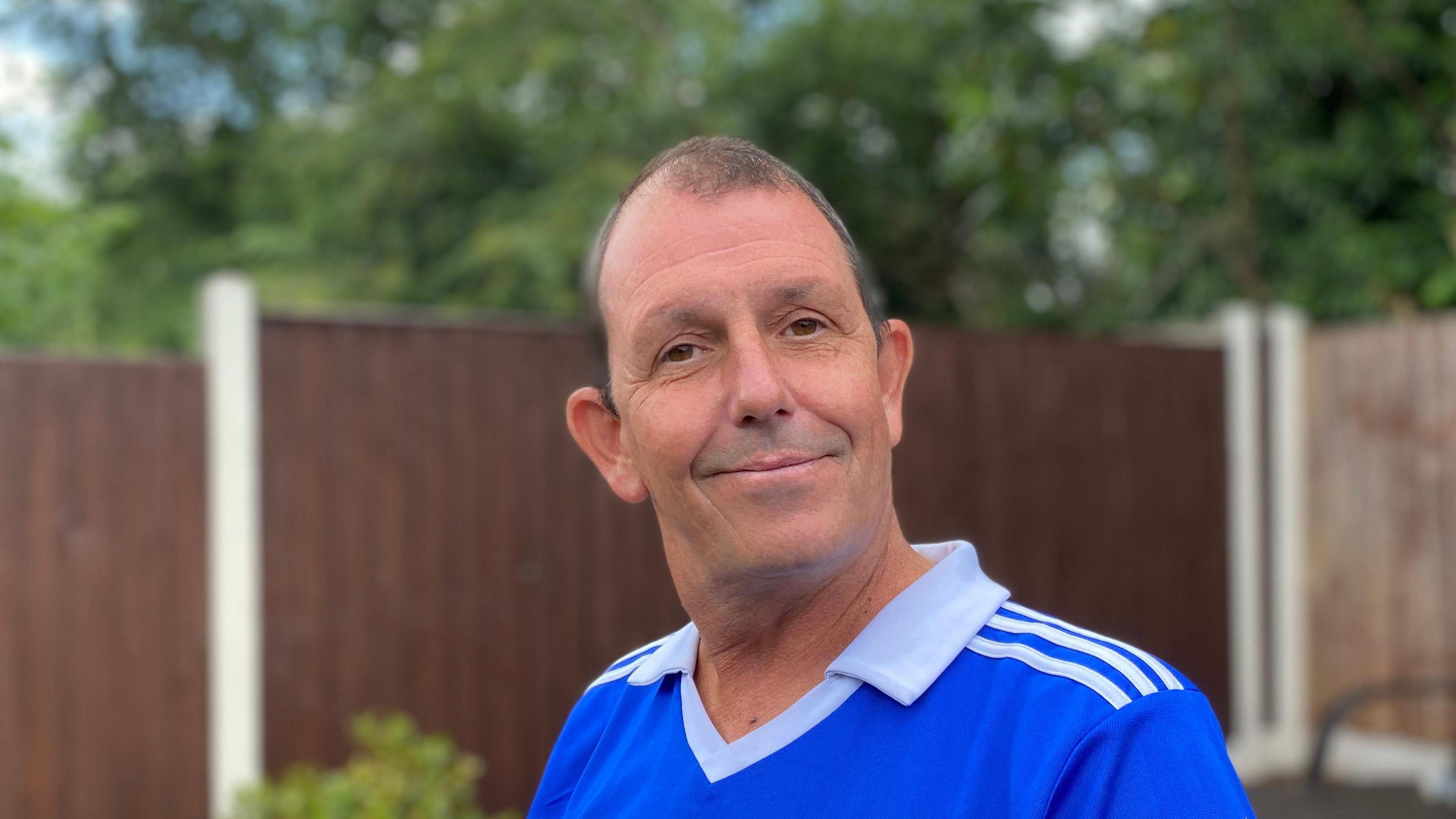 A gentleman, with a blue t-shirt and white collar, poses for the camera in the back garden.