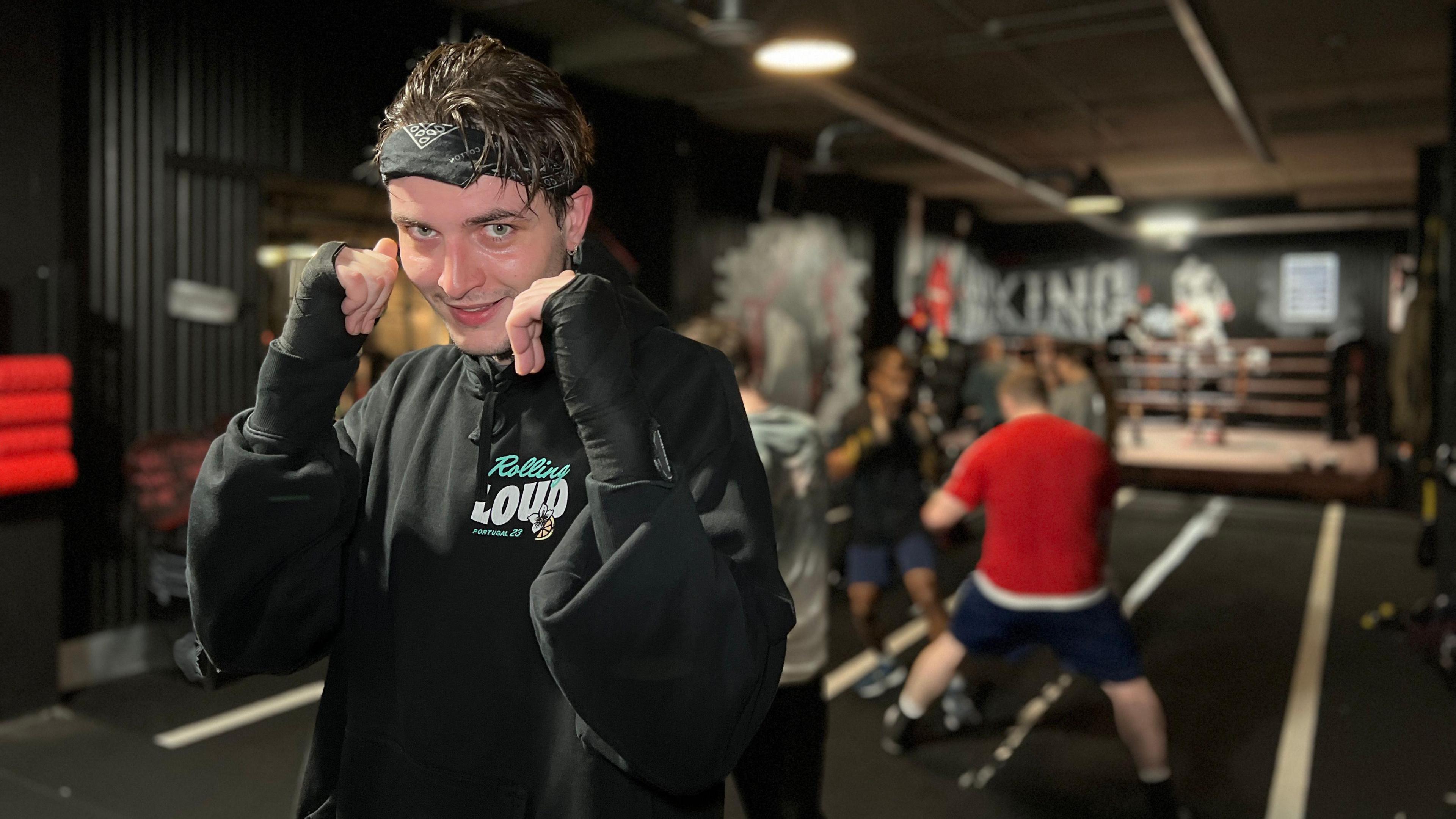 Harry smiles at the camera with his hands up in a boxing pose. His hands are wrapped and he is wearing a black hoodie and a black bandana. Harry has been boxing and behind him people are training.