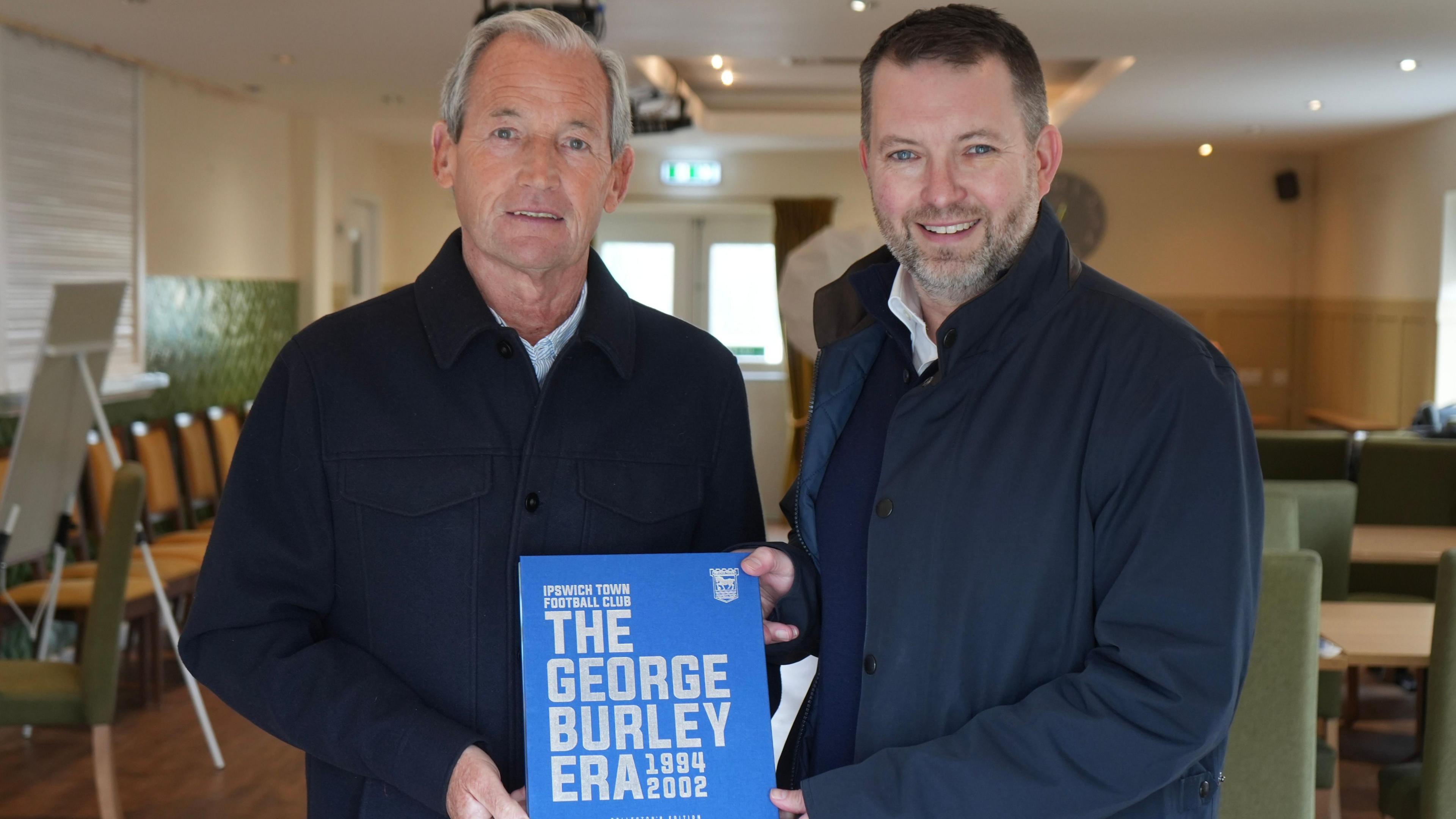 George Burley and Neil Prentice are standing next to each other holding a blue book together that says 'The George Burley Era' in silver writing. Burley is on the left and has grey hair, and is wearing a black coat. On the right is Mr Prentice who has short brown hair and is wearing a navy blue jacket.