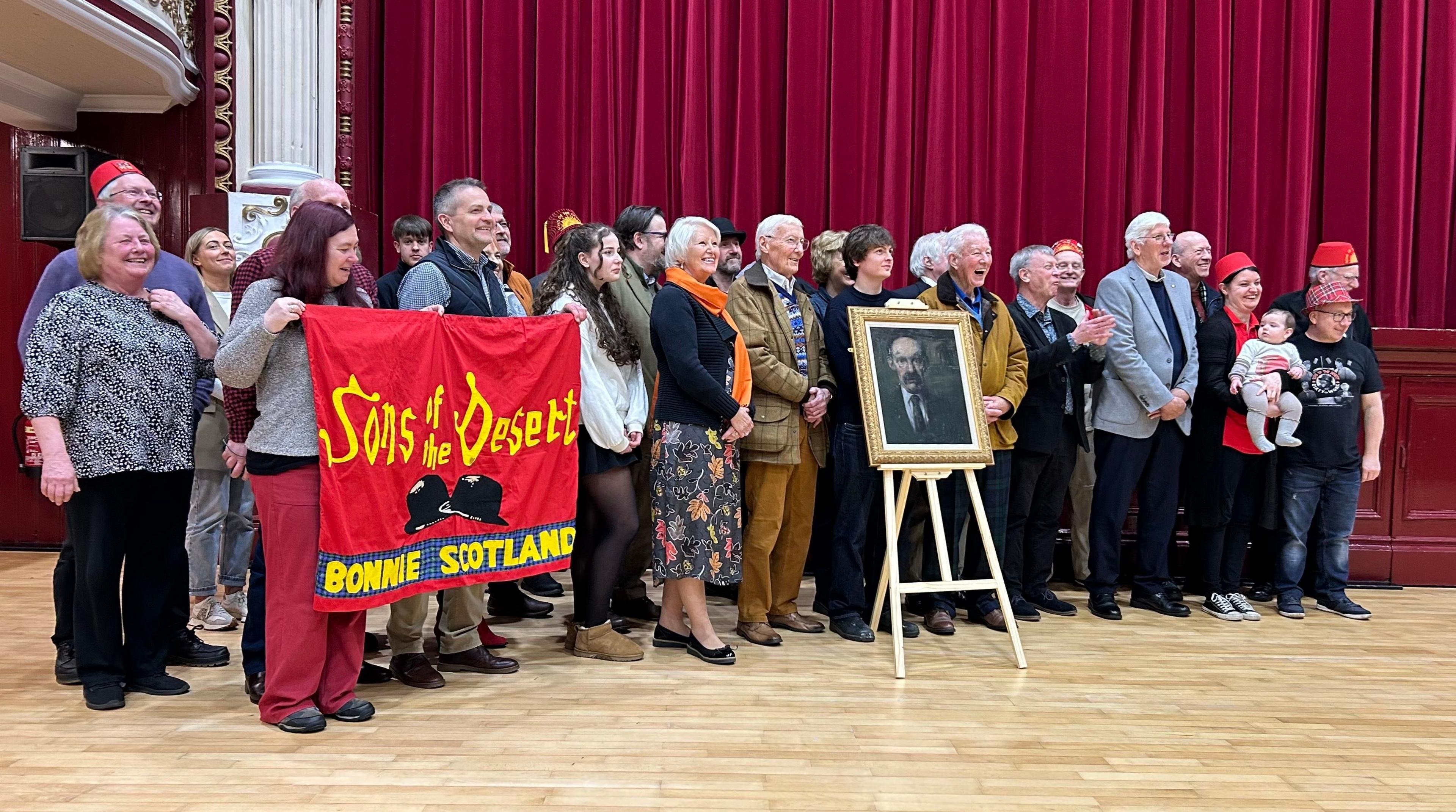 James Finlayson's family and fans gather around a new framed portrait of the actor in front of a stage with red curtains. The oil painting is mounted  on an artist's easel. Two people are holding up a banner showing Laurel and Hardy's famous bowler hats and the words Sons of the Desert, Bonnie Scotland. 