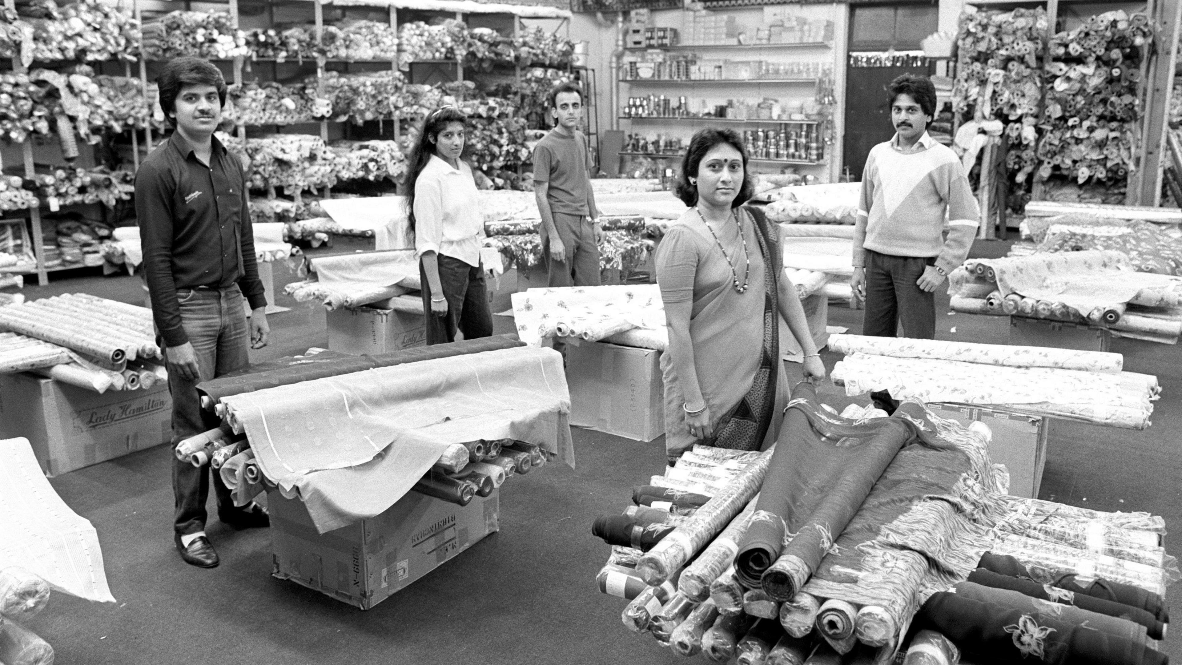 Black and white archive photo of employees of Bombay Store in the 70s surrounded by roles of fabric.