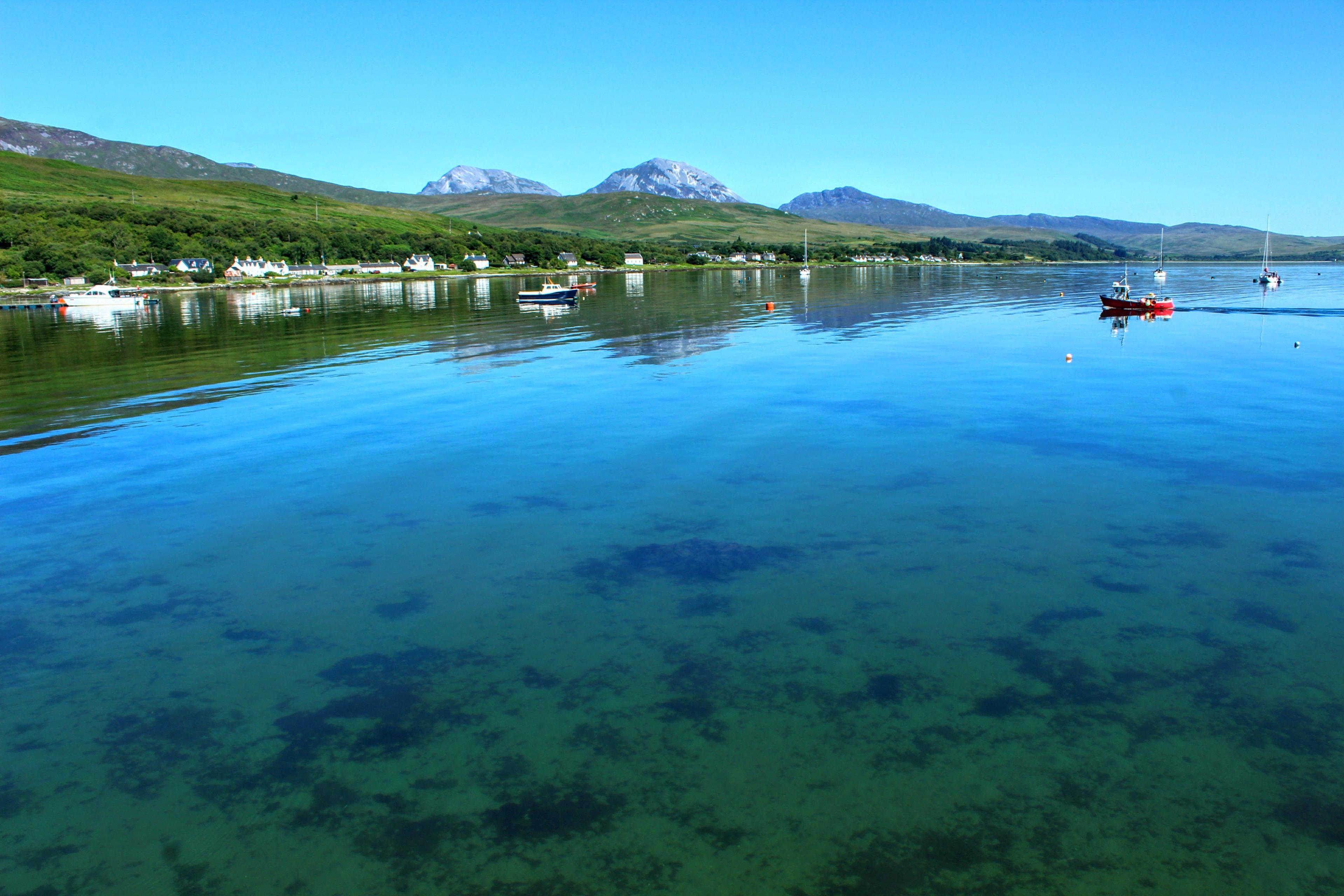 Clear water across the sea on a sunny day, with three mountains and greenery visible behind them 