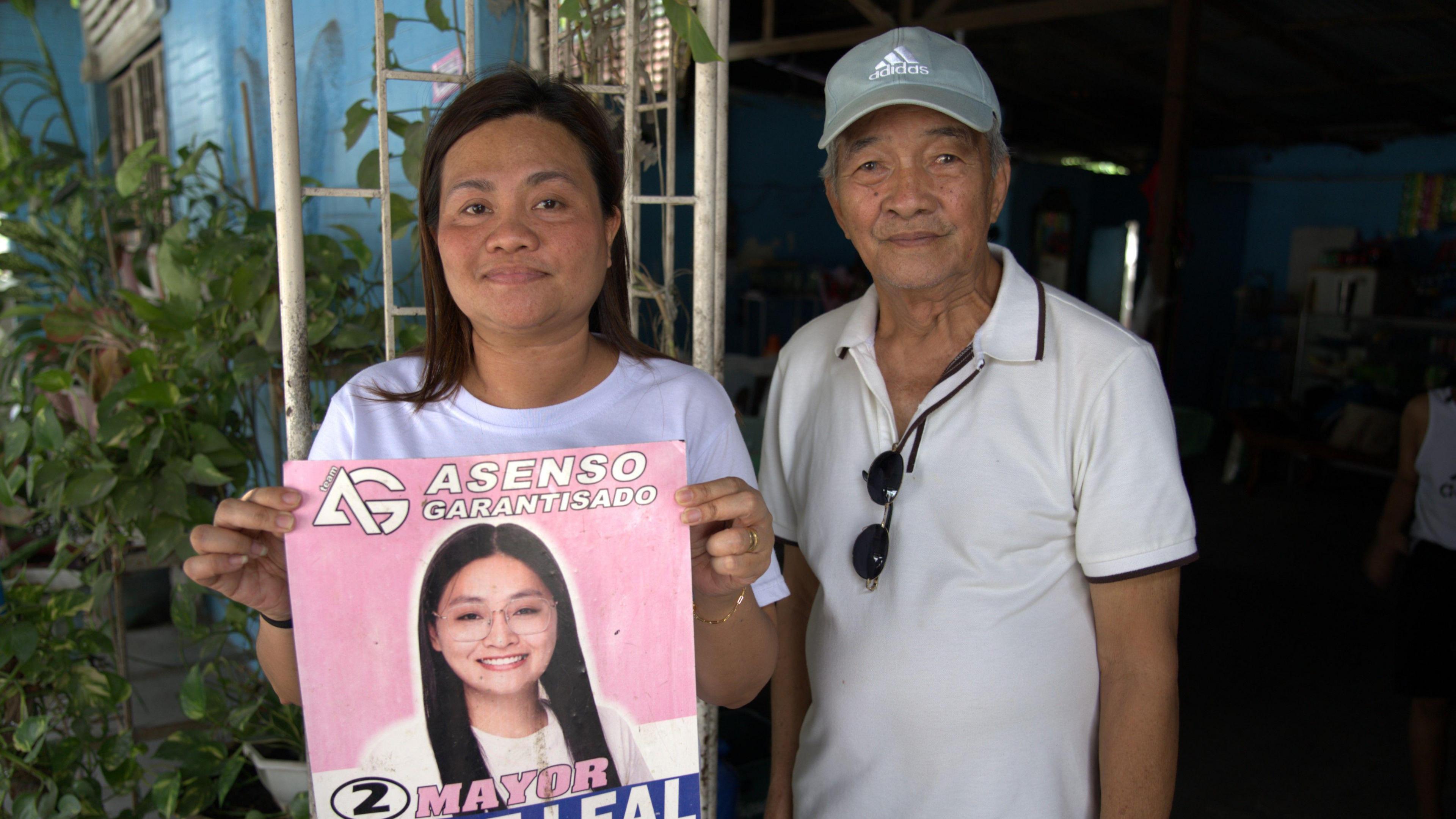 A lady, Miah, holding a pink and blue poster featuring a headshot of Alice Guo with a wide smile above her name in large, white letters. Next to her is an older man, Francisco, wearing a white polo shirt and baseball cap.