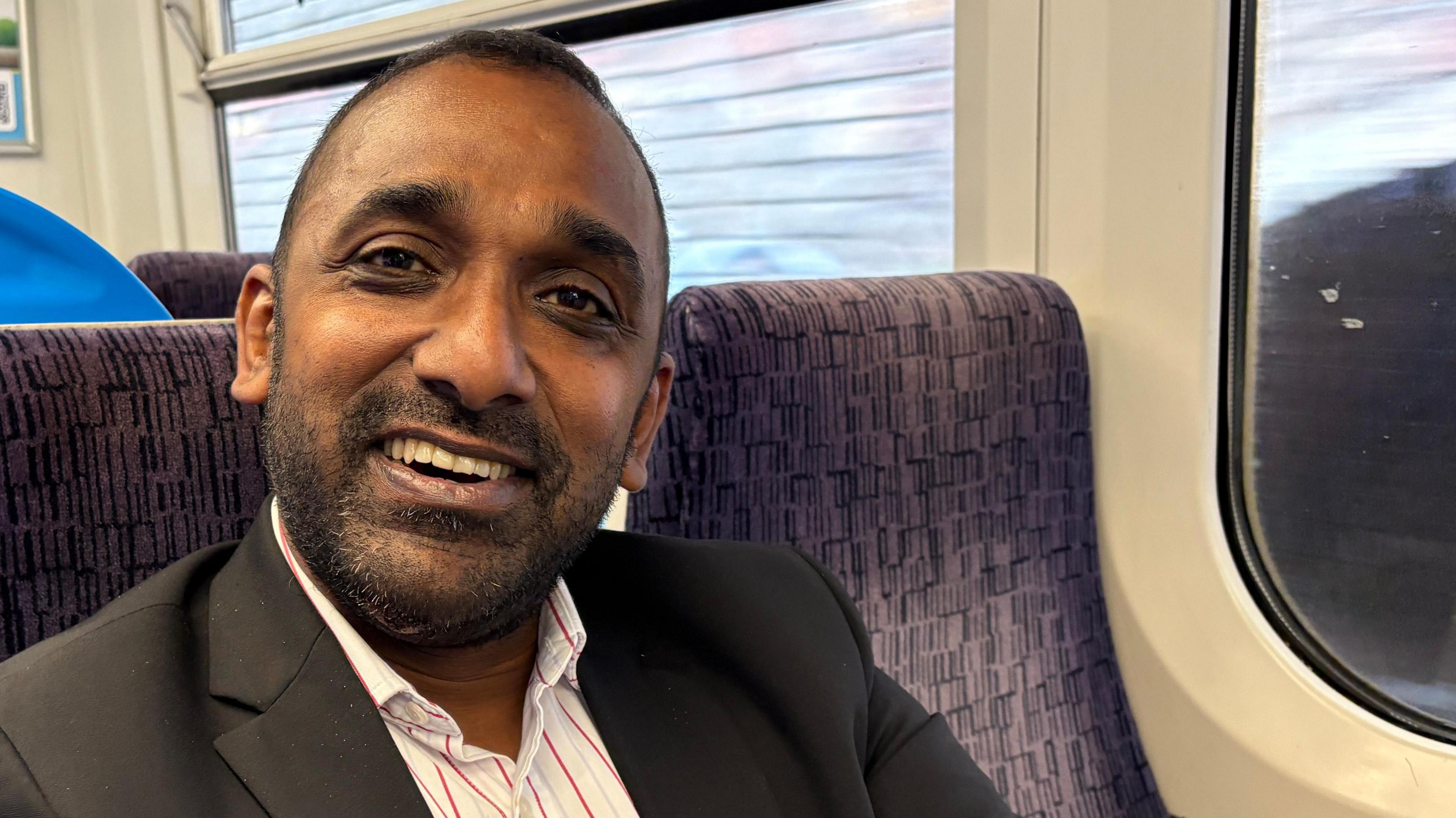 Arvind Kannan, smiling and looking down the camera. He is seated in a train carriage, though the seat next to him is empty. He is wearing an open-necked white shirt with narrow yellow and red vertical stripes, and a black jacket. He has a short beard and shortly cropped hair. 
