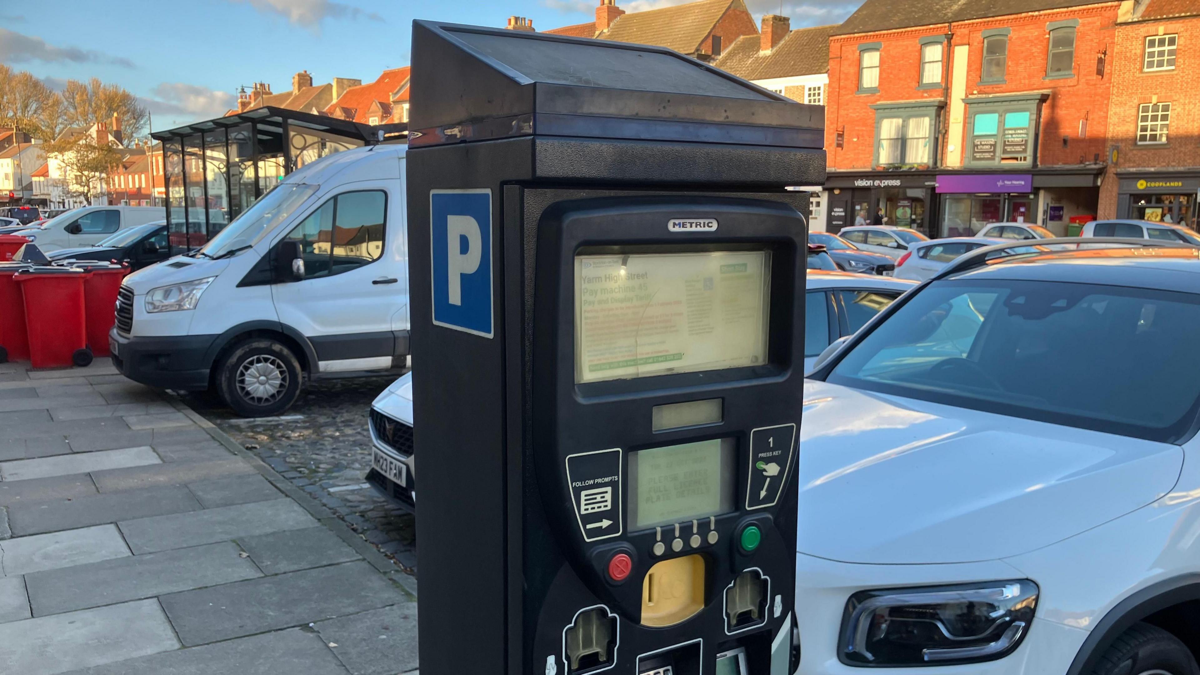 A parking ticket machine in the foreground with Yarm High Street in the background. Lots of vehicles are parked.