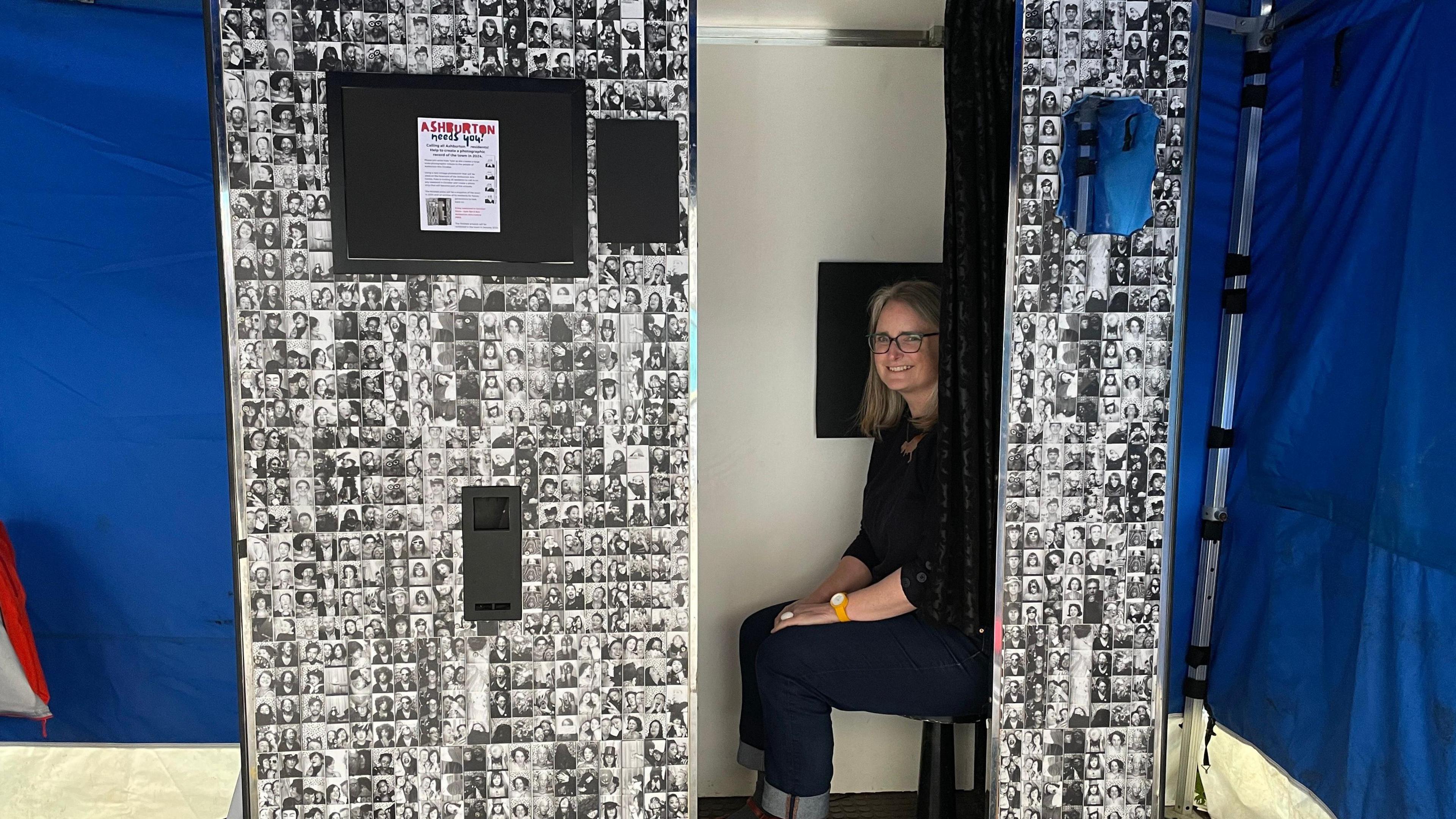 Artist Kate Tyler sits on the chair inside a photobooth with lots of black and white photos of people's faces stuck to the outside 