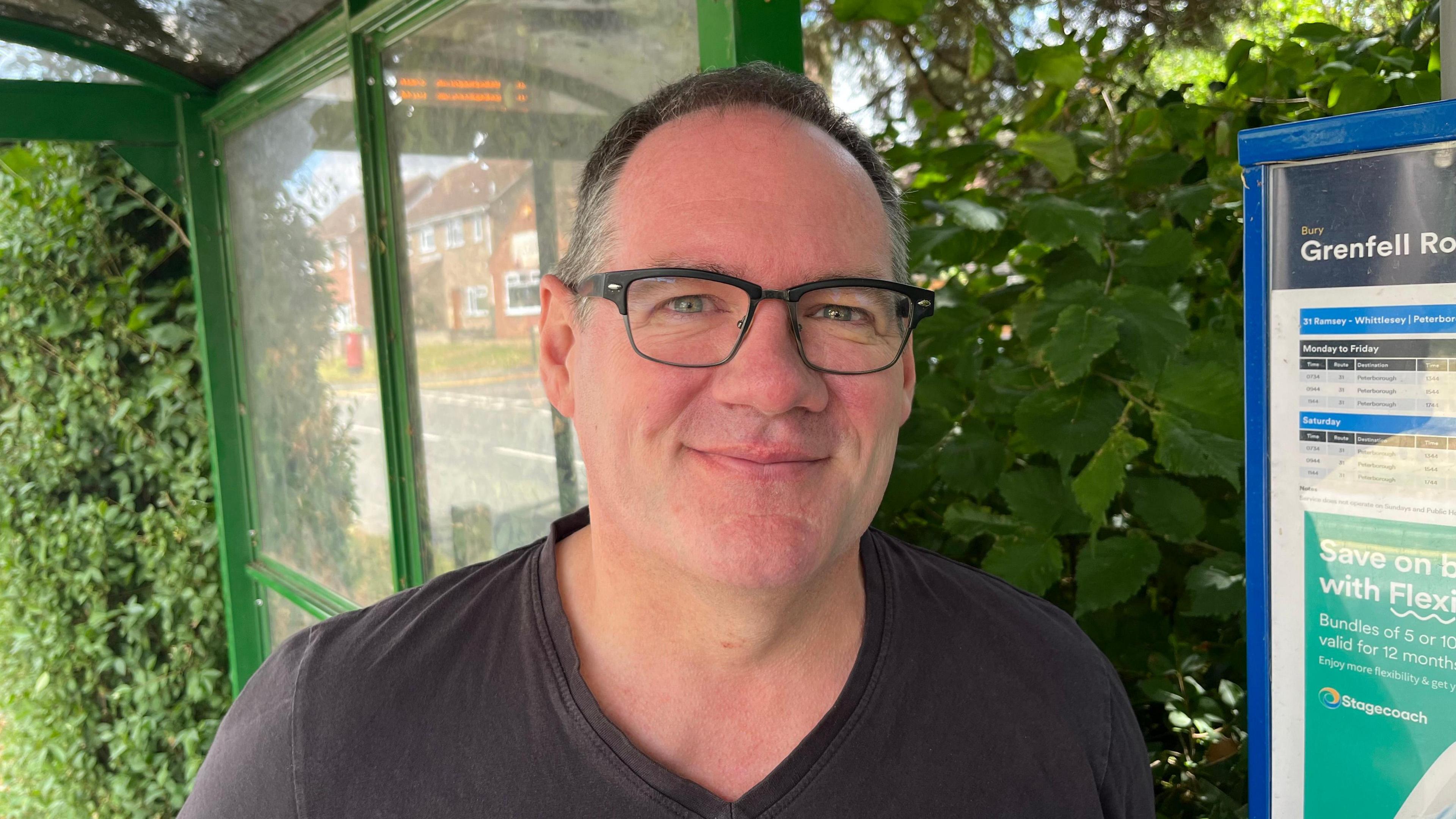 Andrew Copley has dark short hair, with grey flecks, he is wearing glasses with black frames and a dark V-necked T-shirt. Behind him is a bus stop shelter with timetable sign to the right.