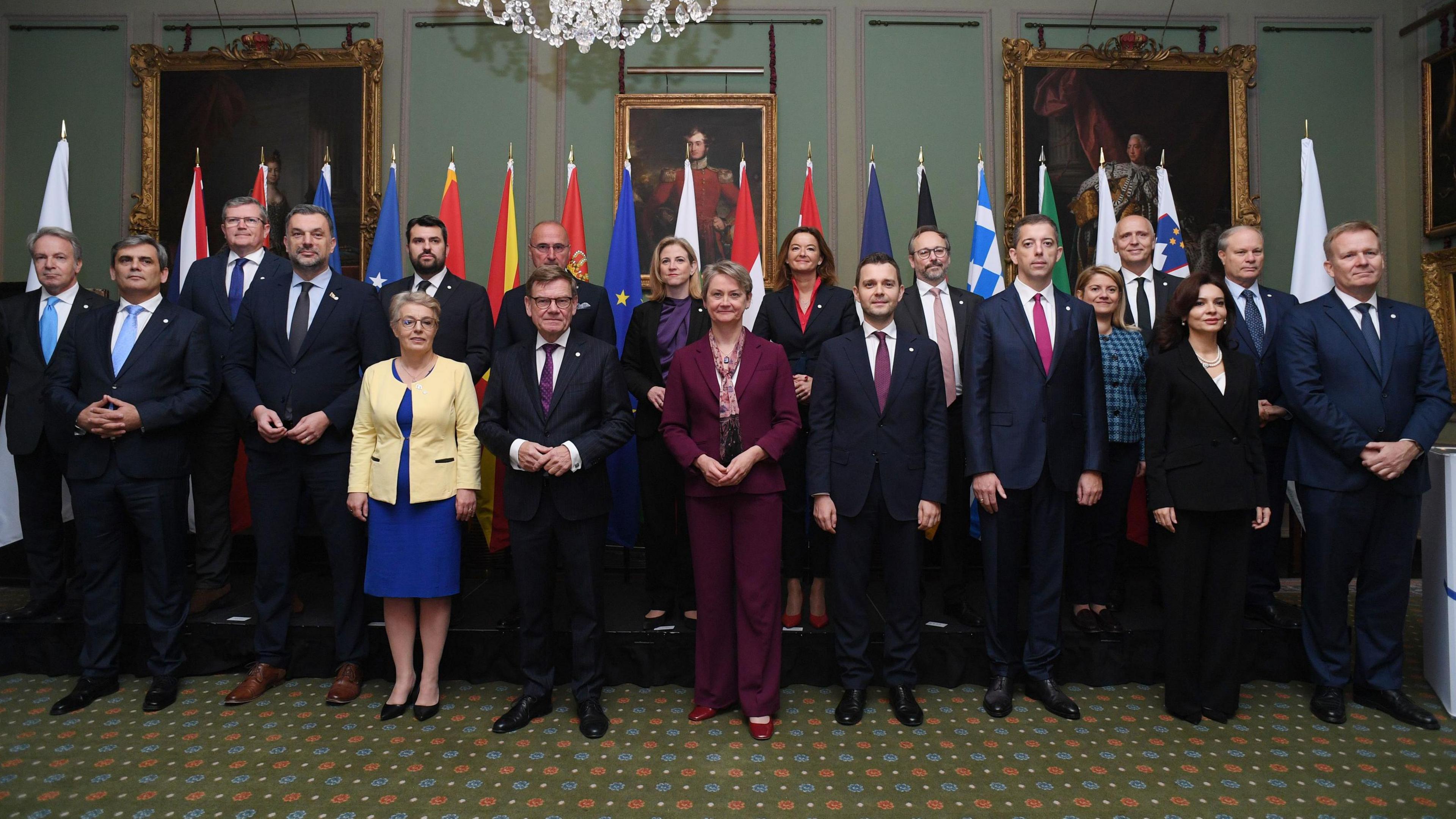 Foreign Secretary Yvette Cooper, who is centre in the front row, poses for a group photograph with other Foreign ministers during the Western Balkans Summit: Foreign Ministers' Meeting at Hillsborough Castle. Cooper is wearing a burgundy suit and is clasping her hands together. Behind the group of ministers are flags representing the different countries.