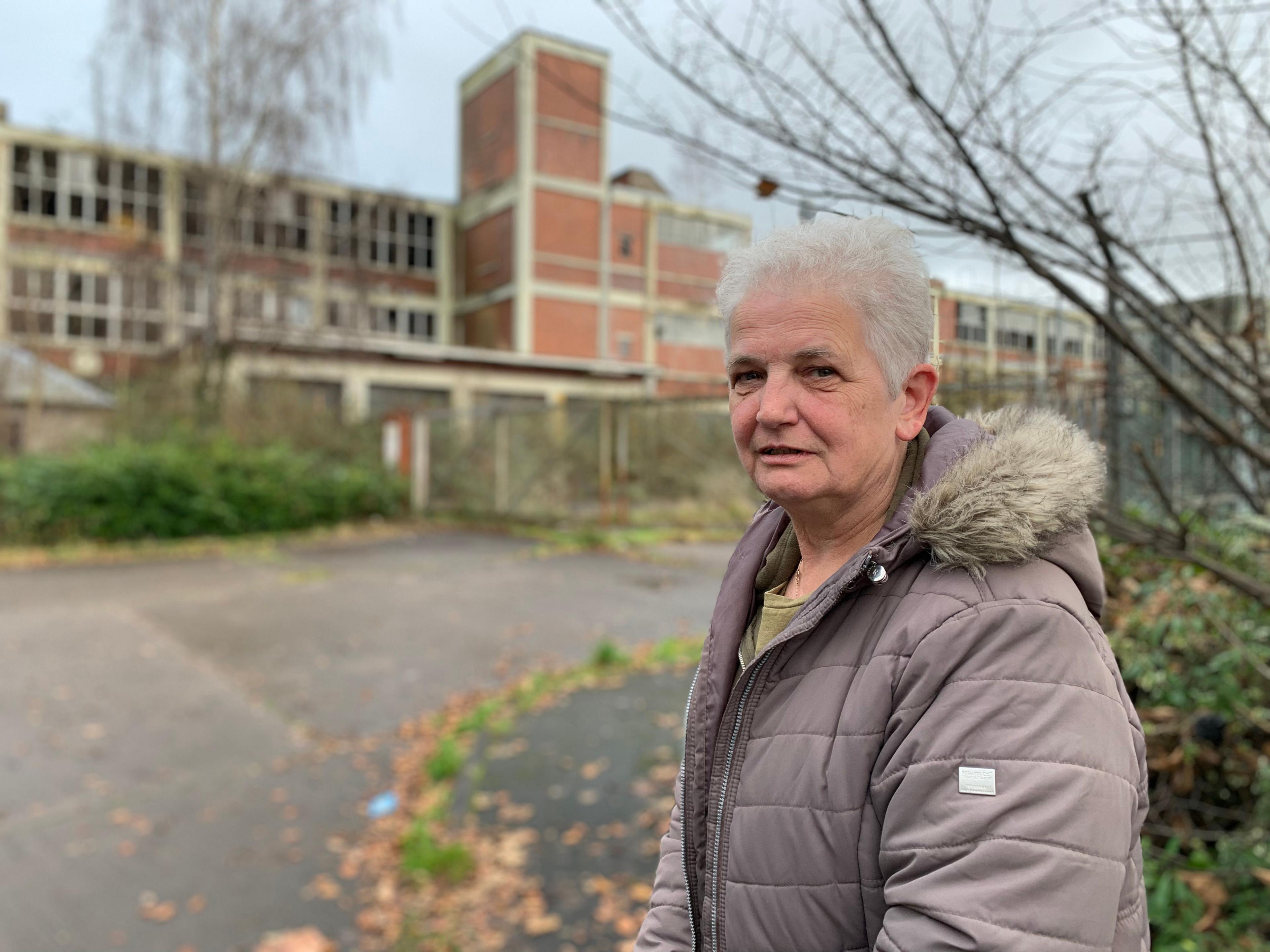 A woman with grey hair in a grey coat stands in front of a factory tower and next to a few tree branches