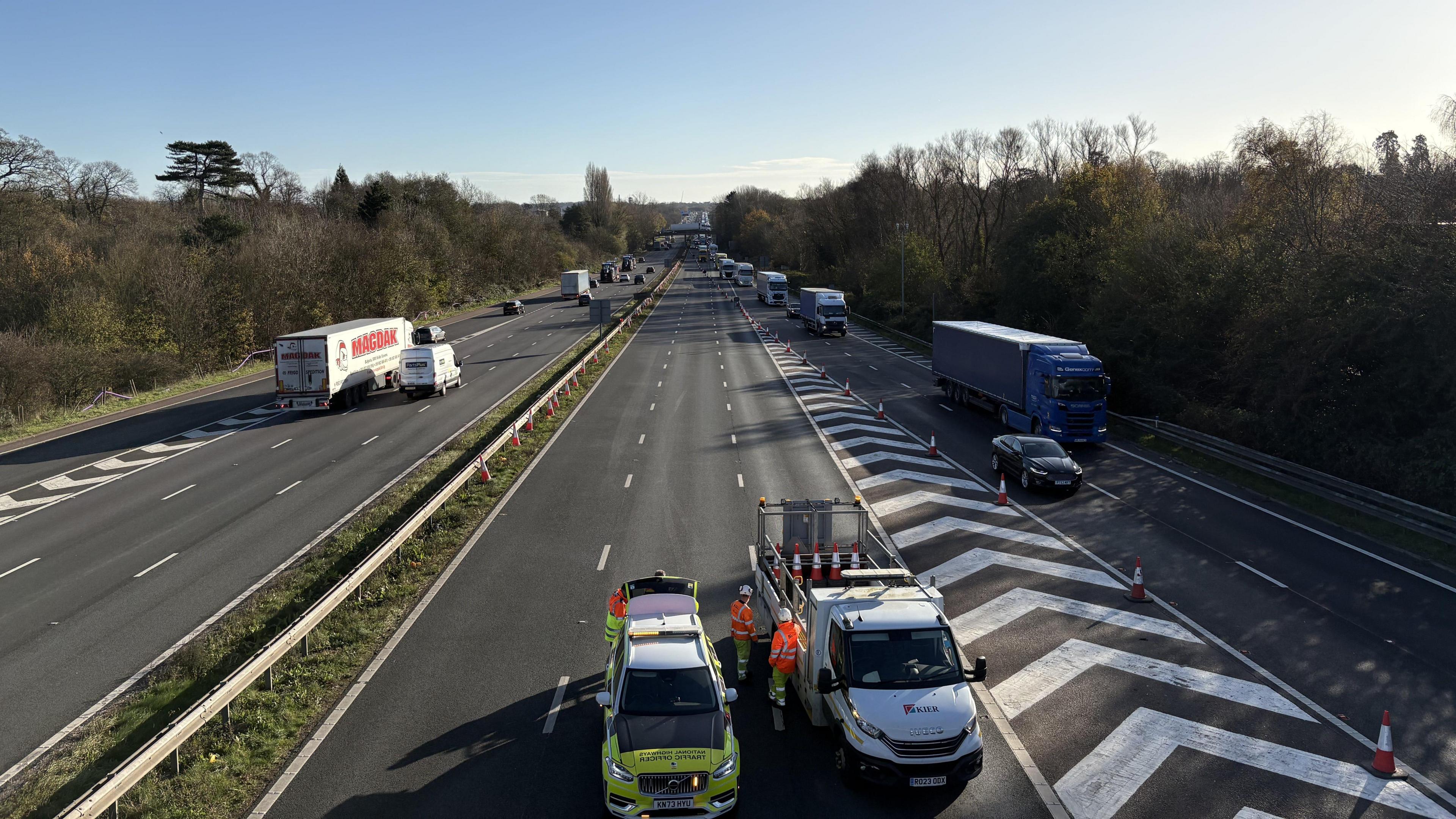 A picture of a motorway from a bridge above. The coastbound carriageway, on the left, is open with traffic flowing. The London-bound carriageway, on the right, is blocked off by Highways Officers and traffic is exiting via a slip road.