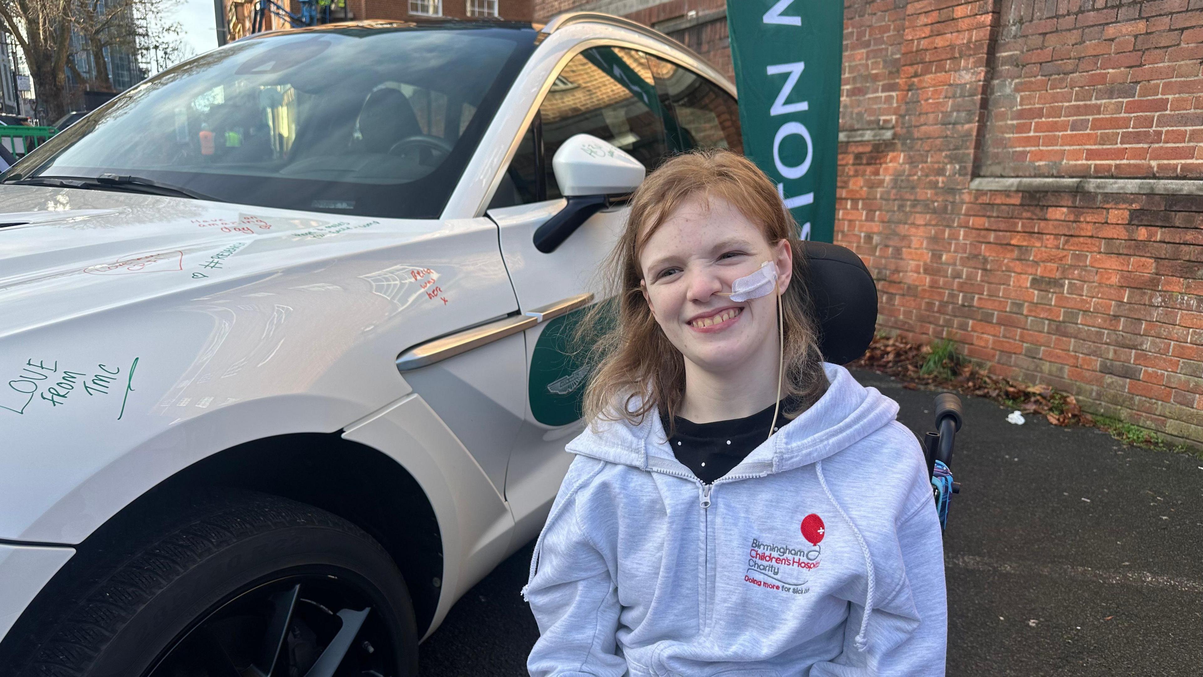 A smiling girl with shoulder length blonde hair in a grey Birmingham Children's Hospital charity hoodie next to a white F1 car in a carpark. She is in a wheel chair and has a tube going into her nose. A brick wall is behind her.