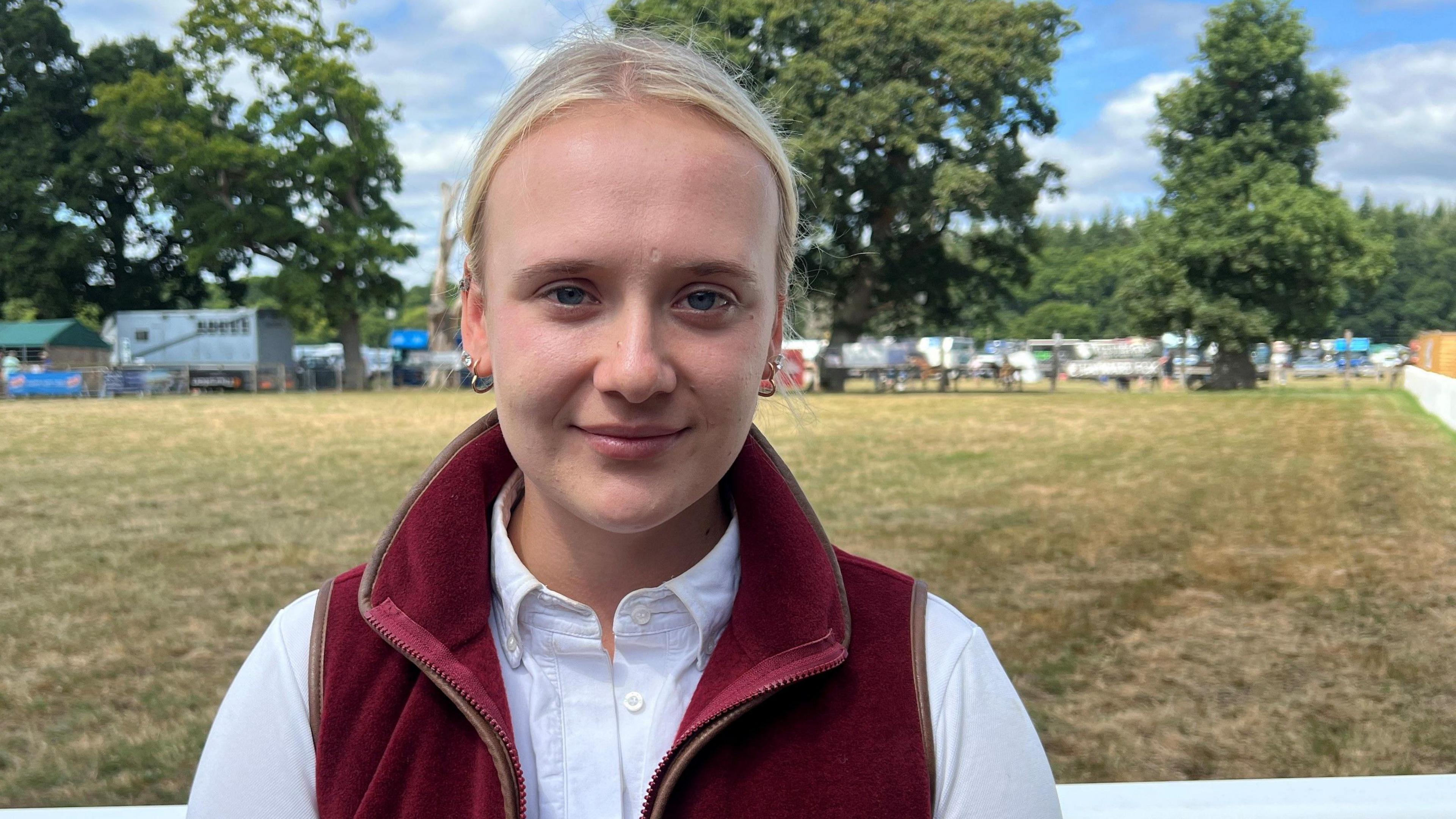 Anna Merritt wearing a white top with sleeveless burgundy fleece, standing in front of an open field at the New Forest Show.