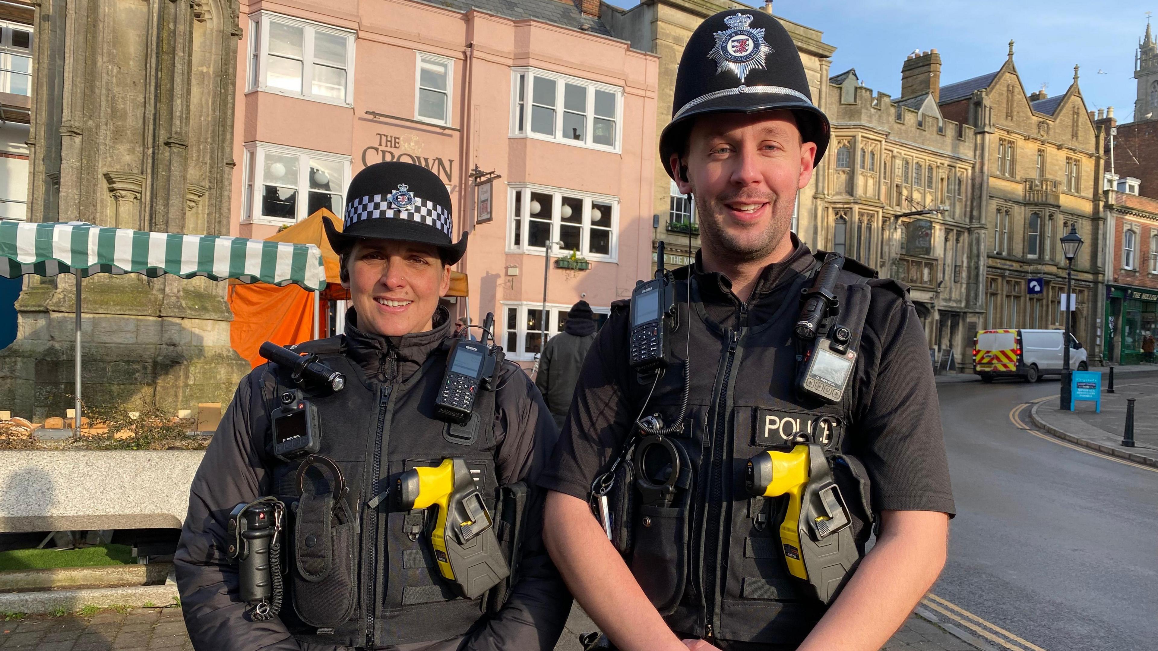 The picture shows PCs Stacey Bennett and James Higgins standing by the Market Cross in Glastonbury.  They are smiling at the camera. It's a sunny day and there is a market stall in the background.