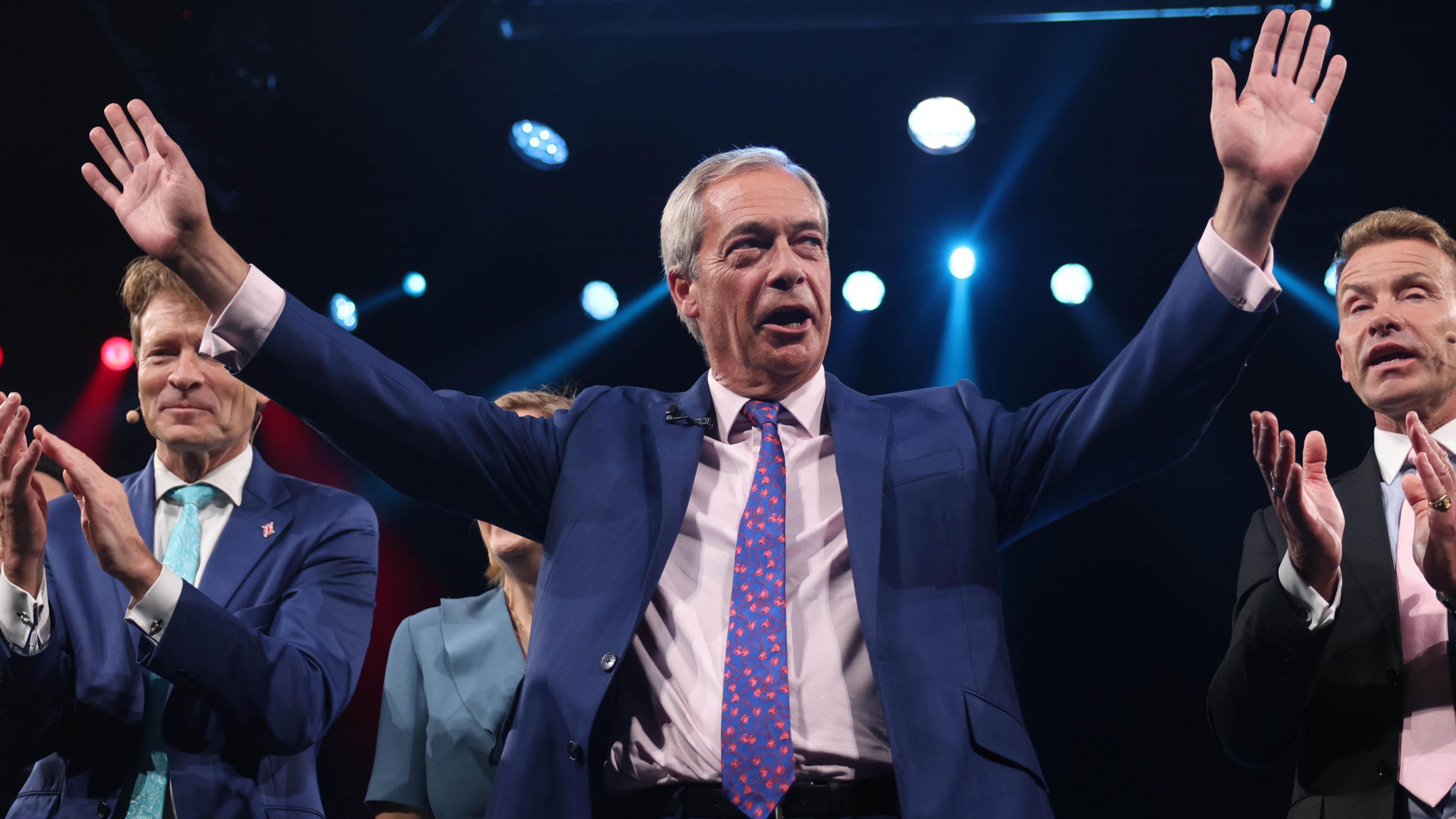 Nigel Farage with his arms raised, acknowledges the crowd at the Reform conference. Beside him, two senior Reform figures applaud.