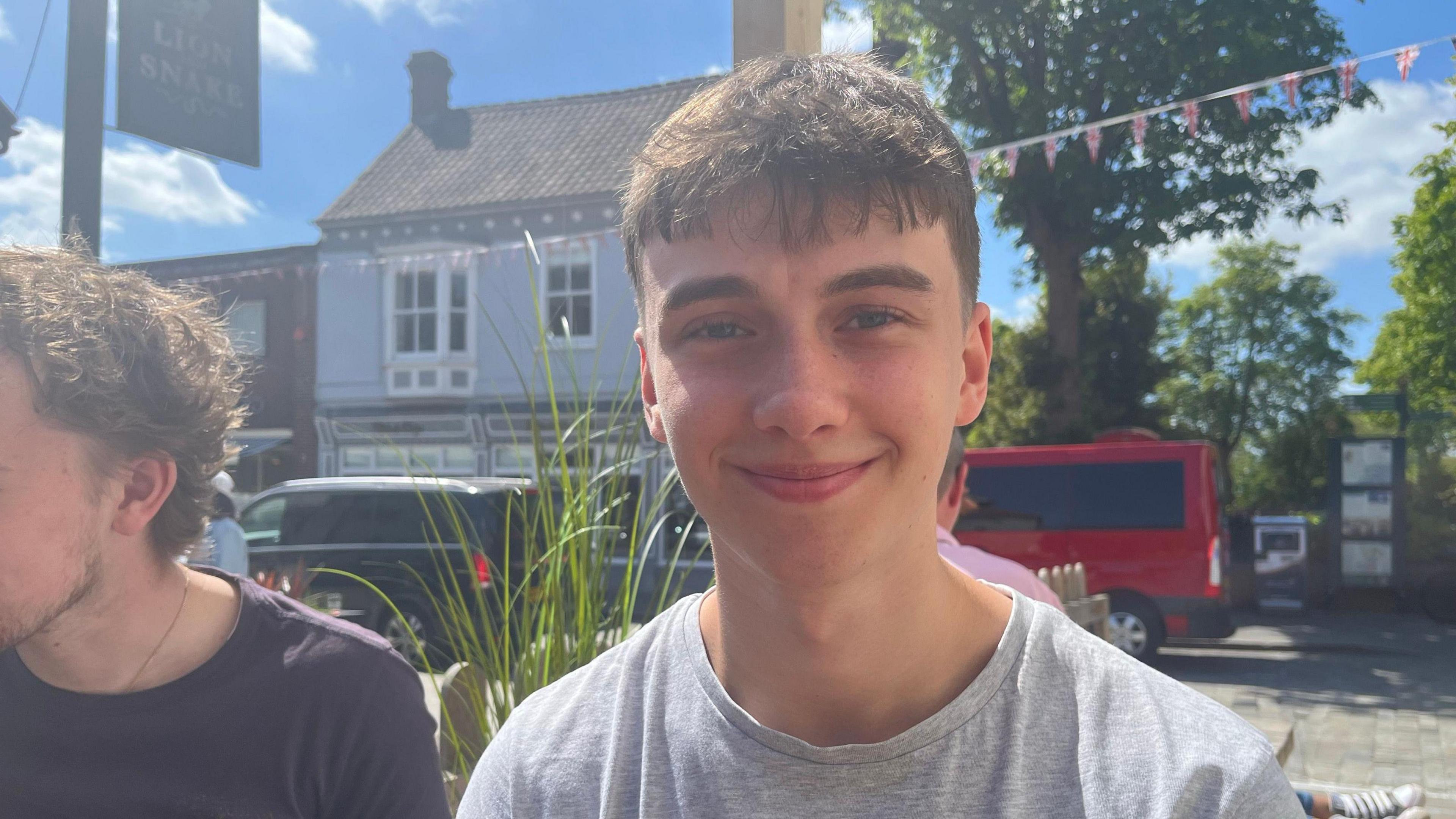 A man smiling at the camera. He is wearing a grey t-shirt. There are buildings and cars parked behind him. The sky is blue.