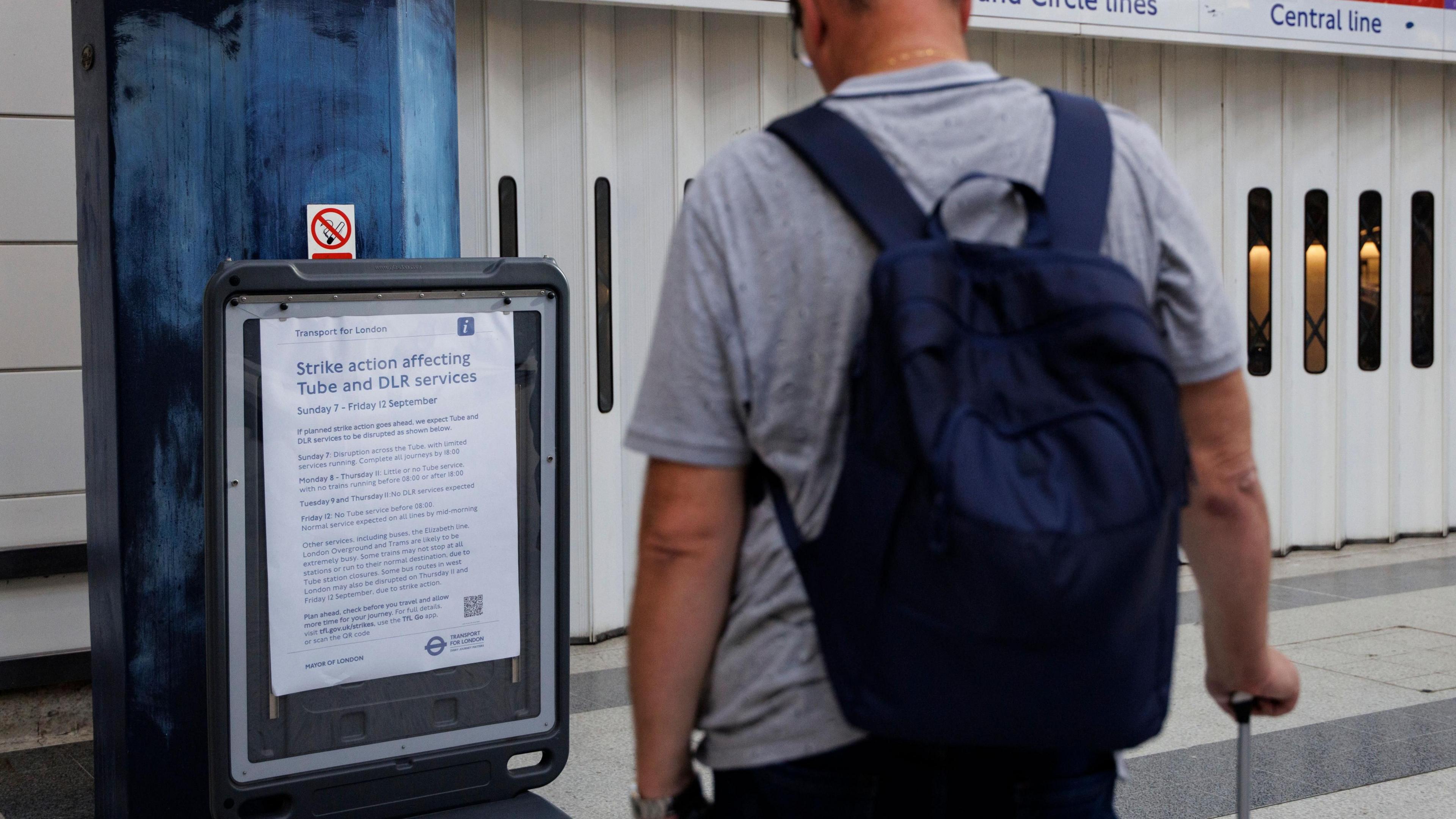 A man stands with his back to the camera looking at an information board. In front of him is the shuttered entrance to a Tube stations. 