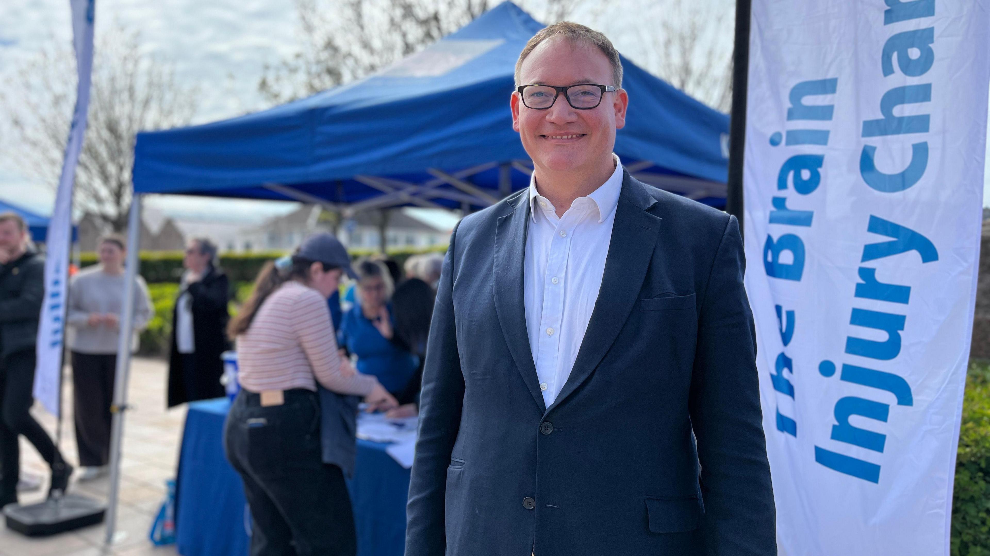 Edward Loader smiles at the camera as he wears a suit and behind him people are checking their pétanque scores at the Headway Jersey stand