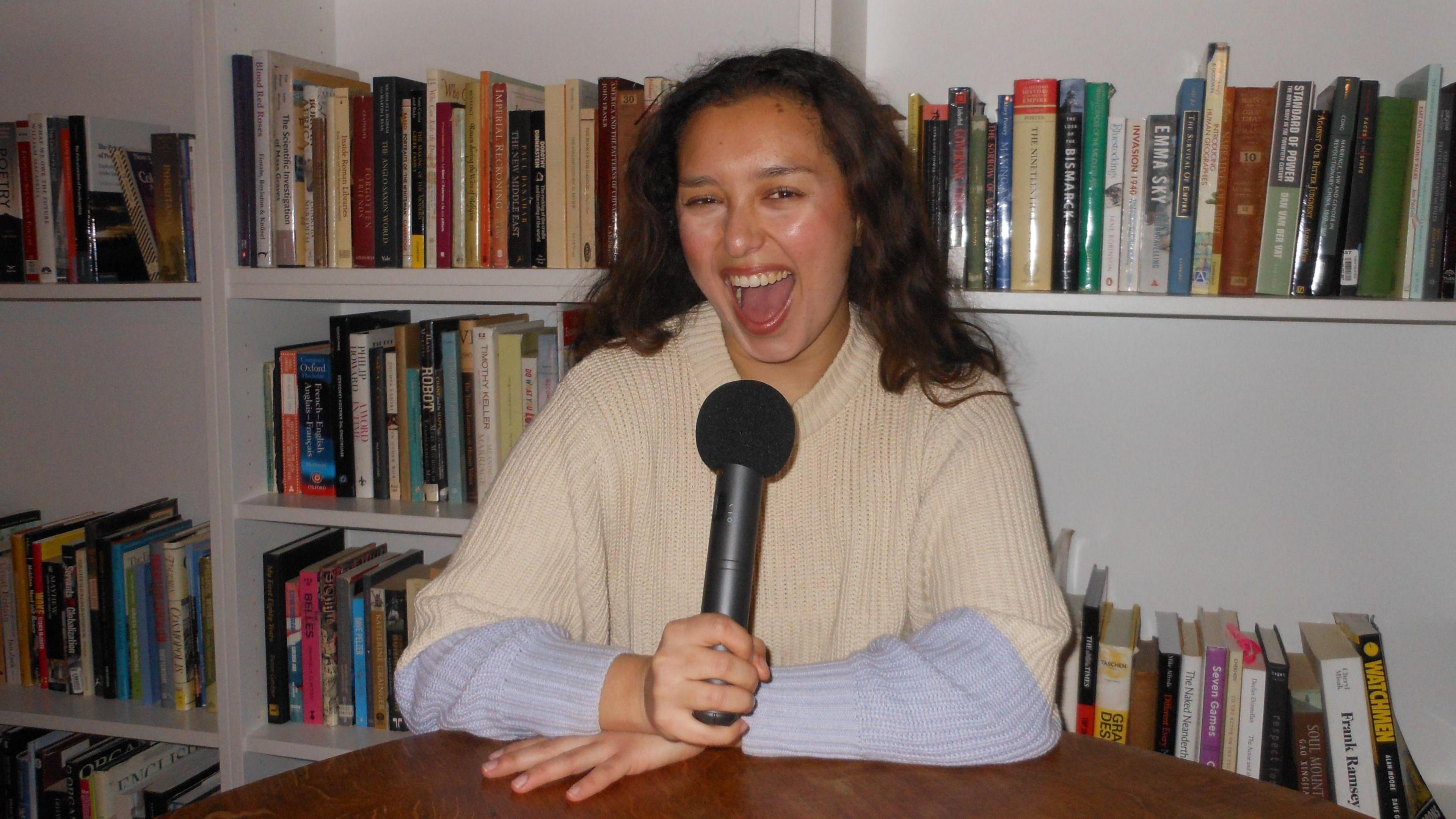 Lara Harris pictured at a table with shelves of books behind her. She has long dark wavy hair and is holding a black microphone.