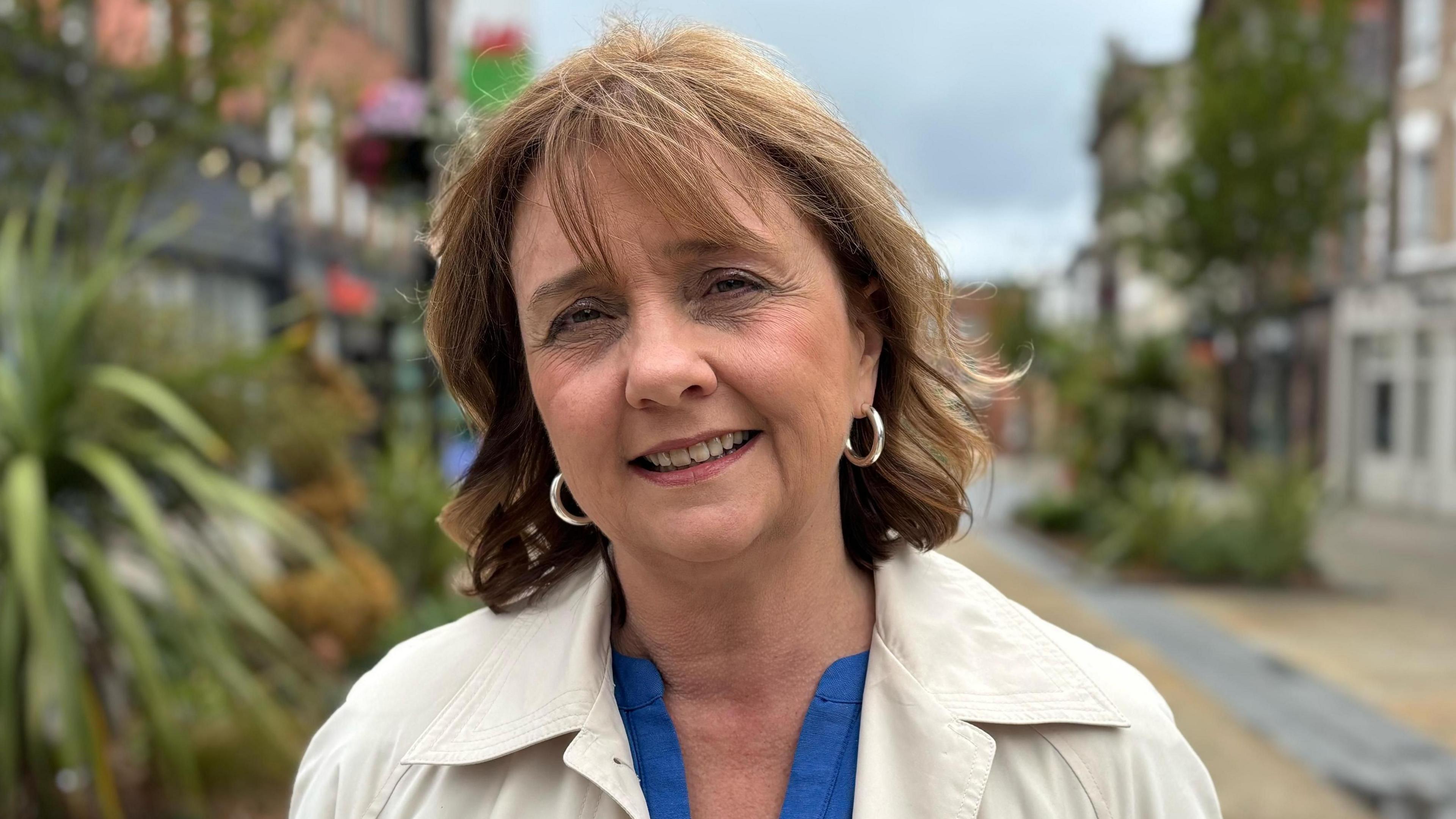 Llinos Roberts smiling at the camera. She has short brown hair and is wearing hoop earrings. She is standing in Wrexham city centre surrounded by potted shrubbery. She has a cream coat with a blue shirt underneath. It is a head and shoulders shot of her.