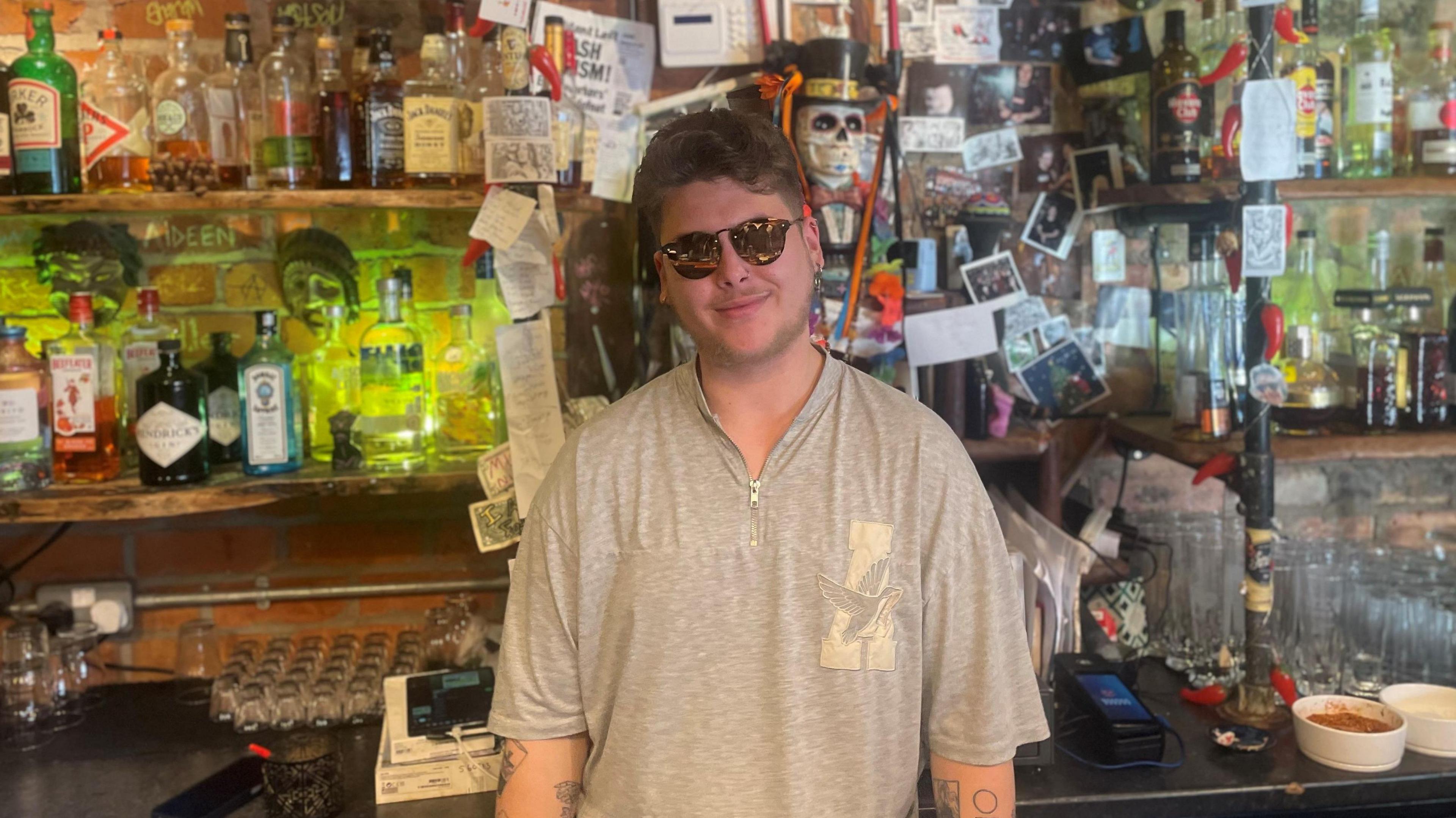 Conor Lynch in a grey top, wearing sunglasses stands behind the bar at a pub. He has short brown hair and a tattoo can be seen on his lower arm.