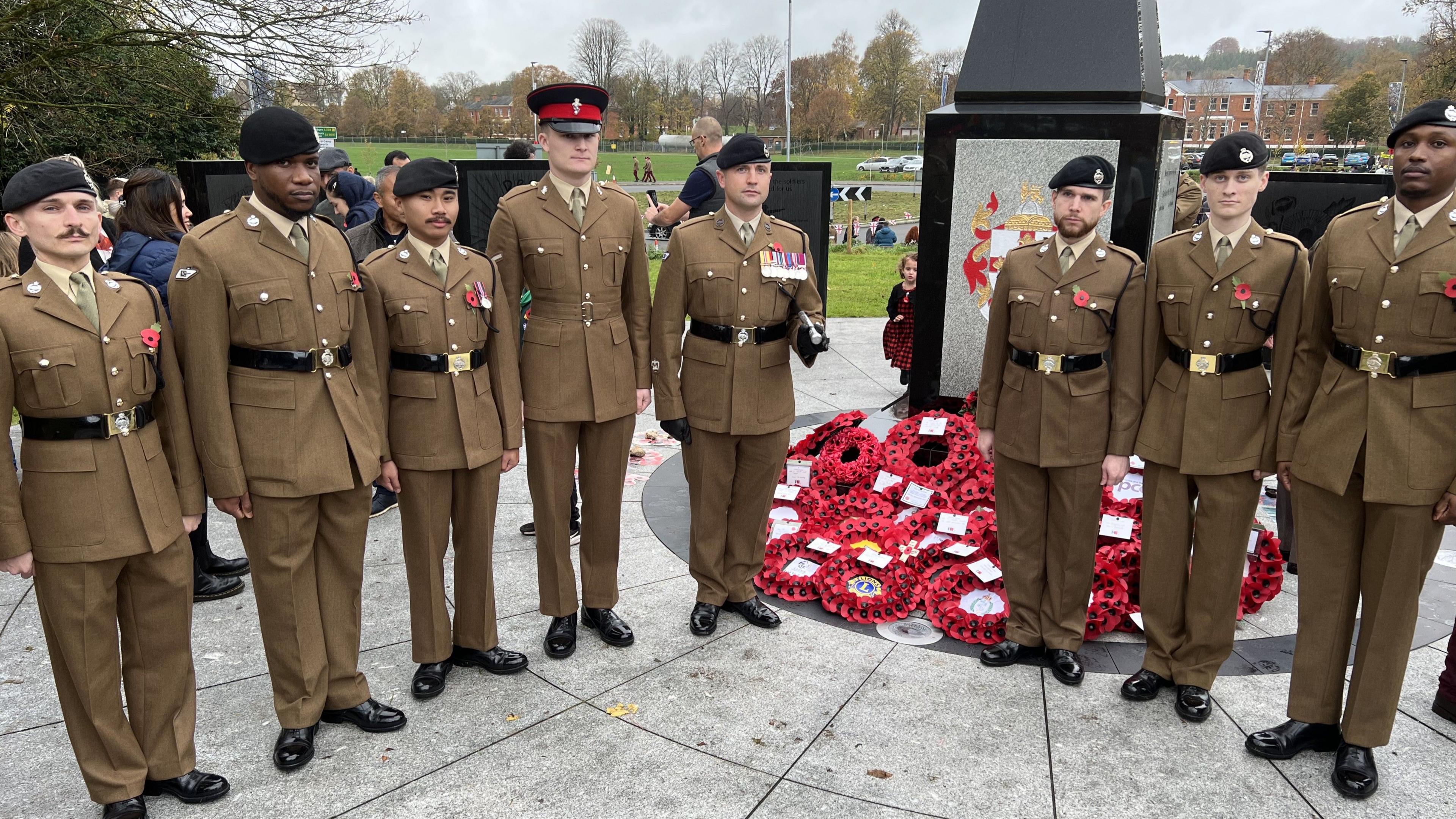 A line of eight servicemen wearing brown uniforms and black belts, standing to attention beside a cenotaph and posing for a picture. There are lots of poppy wreaths laying at its base.