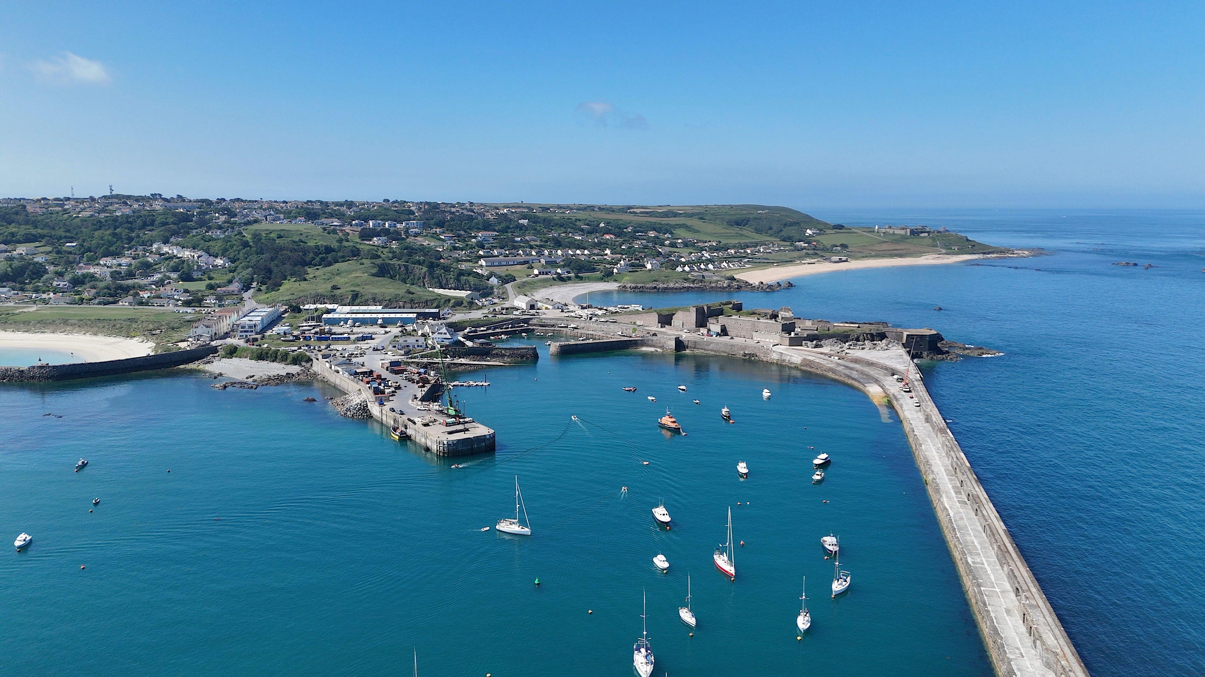 The harbour in Alderney from the air. The long breakwater is reaching out of the photo and nestled in the harbour is a number of boats with sails including the Alderney lifeboat.